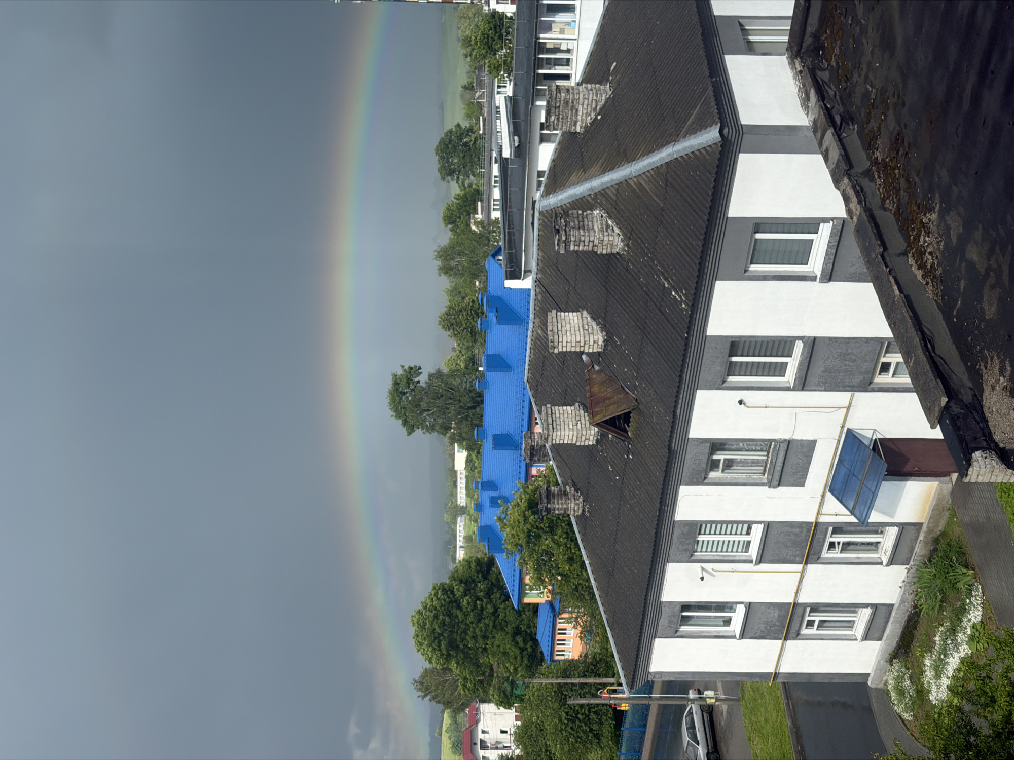An aerial view captures a residential area in Valozhyn, Belarus, under a dramatic sky with a rainbow. In the foreground, a two-story building with a dark, corrugated roof is prominent. This building has a distinctive facade with alternating vertical bands of white and gray. It features multiple white-framed windows, some with blinds. A small blue awning shelters a brown door on the right side of the building.
Further back, a row of buildings with bright blue roofs suggests a communal or institutional setting. These buildings are interspersed with lush green trees. In the distance, more buildings and rolling, green hills are visible under a dark, overcast sky, partially obscured by mist or rain. A faint rainbow arcs across the sky, spanning from the left to the right of the frame, hinting at recent or ongoing precipitation. A silver car is parked on the wet asphalt road to the left of the foreground building. The overall scene depicts a somewhat subdued urban landscape, illuminated by the striking natural phenomenon of a rainbow.