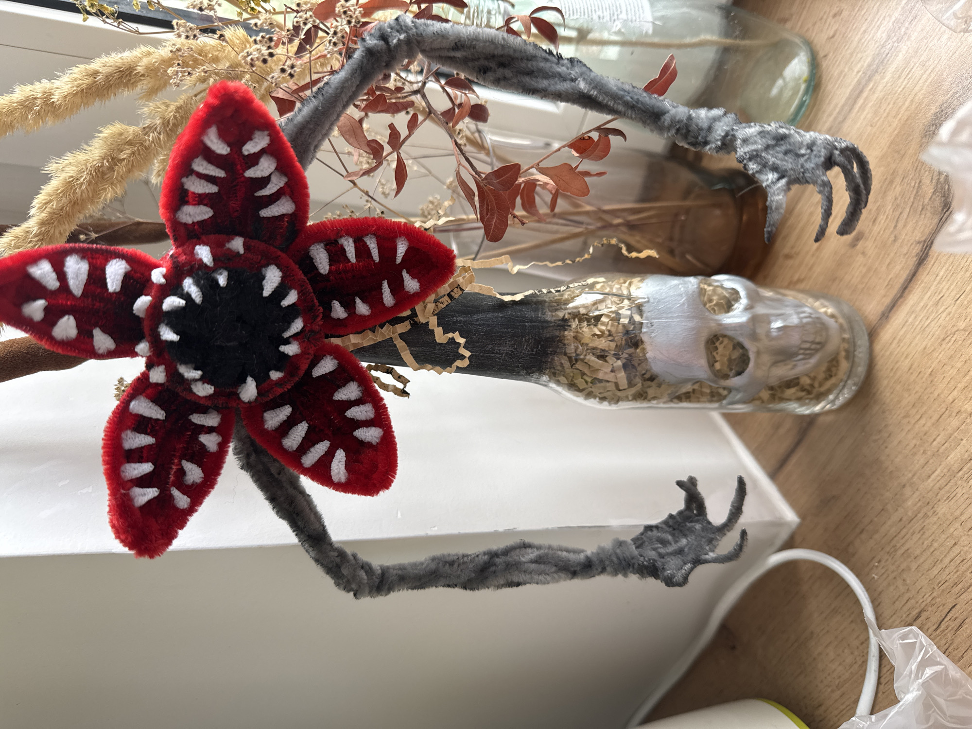 In an indoor setting, possibly a home or studio, a crafted effigy resembling a Demogorgon from Stranger Things is the central focus. This effigy is constructed from red and black pipe cleaners, with white pipe cleaners forming sharp teeth. Its body appears to be a glass bottle, painted silver to resemble a skull at its base, and filled with shredded brown material. Two grey pipe cleaner arms extend from the creature's head, each ending in claw-like hands. The effigy is positioned against a backdrop that includes a white surface, possibly a wall or curtain, and a collection of dried floral arrangements in glass vases. The foreground shows a wooden surface with a white electrical cord and a partially visible plastic bag. The lighting suggests natural light is coming from the right.