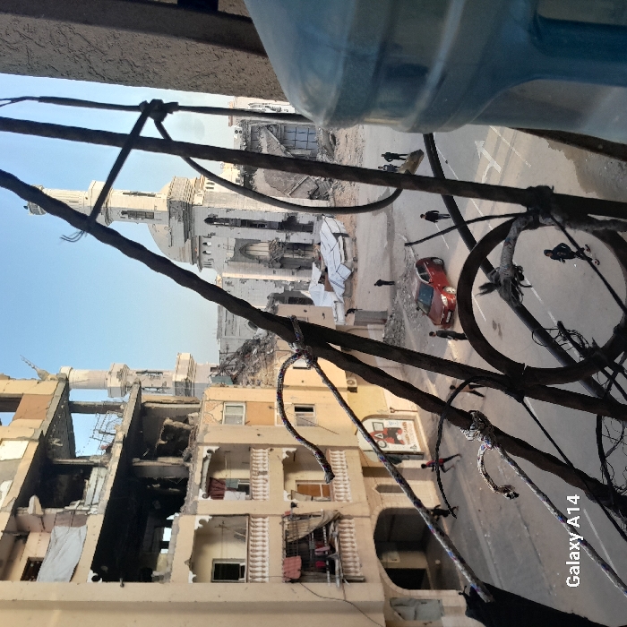 A scene of urban devastation is visible through a tangle of wires and ropes. Buildings are heavily damaged, with exposed interiors and rubble. A mosque stands amidst the destruction. On a road below, a red car is visible, and several individuals are walking. The image is shot from an elevated perspective, partially obscured by a large plastic water bottle in the upper part. "Galaxy A14" is visible in the lower right.