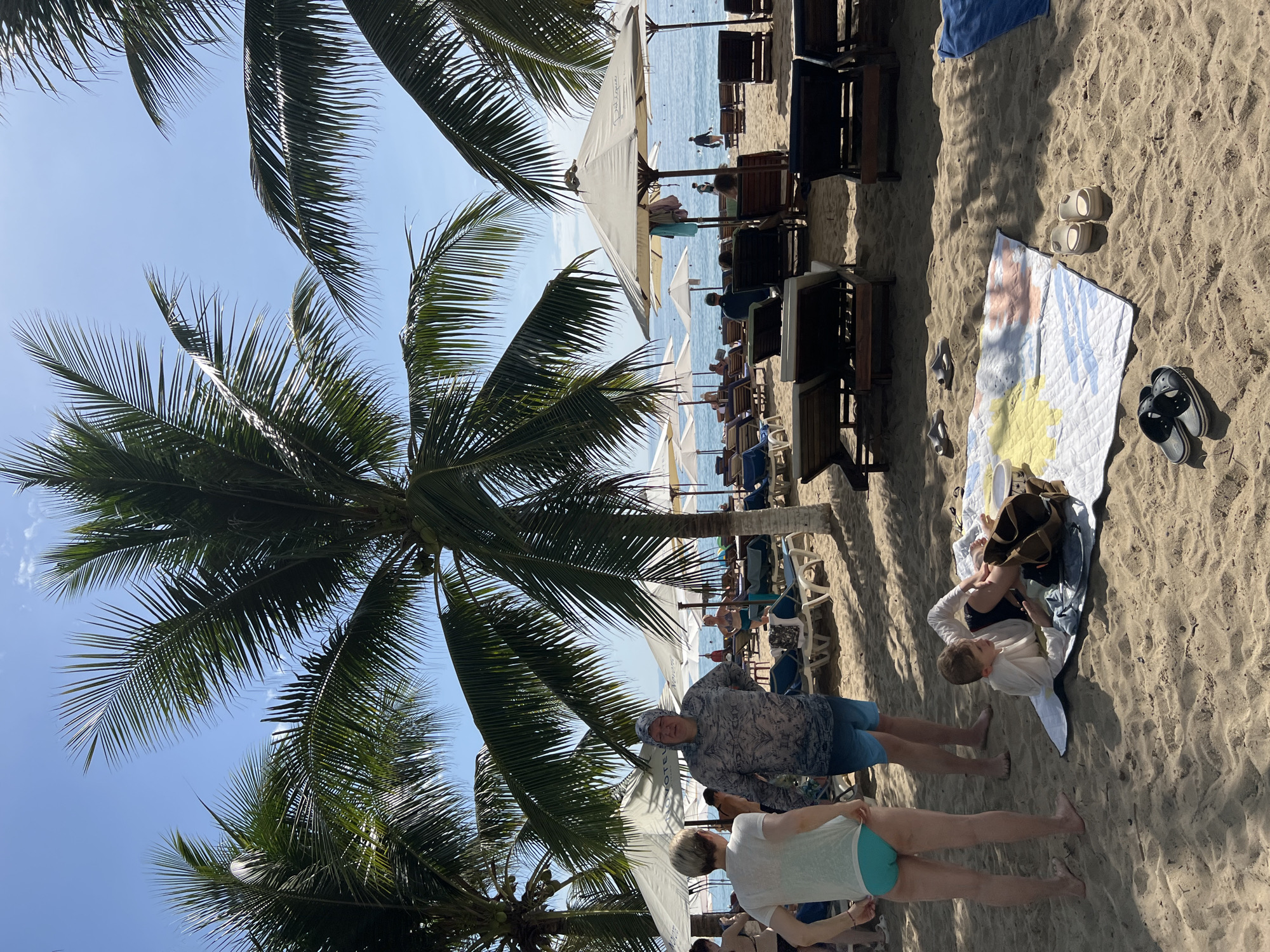 The image depicts a sunny day at a beach in Nha Trang, Vietnam. In the foreground, a young boy lies on a colorful blanket on the sand, appearing to be playing with a phone. Beside him are various footwear, including flip-flops and sandals. A woman in a white t-shirt and turquoise bikini bottoms stands to the left, while a man in a hooded long-sleeved shirt and blue shorts stands to her right.
In the middle ground, rows of empty lounge chairs and umbrellas are set up on the sand, indicating a resort or public beach area. The umbrellas are cream-colored and some have logos visible. Further back, people are seen in the distance, some walking along the shoreline and others relaxing under umbrellas. The ocean stretches to the horizon, with calm, blue water and a clear sky above. Large palm trees with lush green fronds dominate the scene, casting dappled shadows on the sand and framing the view of the beach and sea. The overall impression is one of a relaxed beach environment.