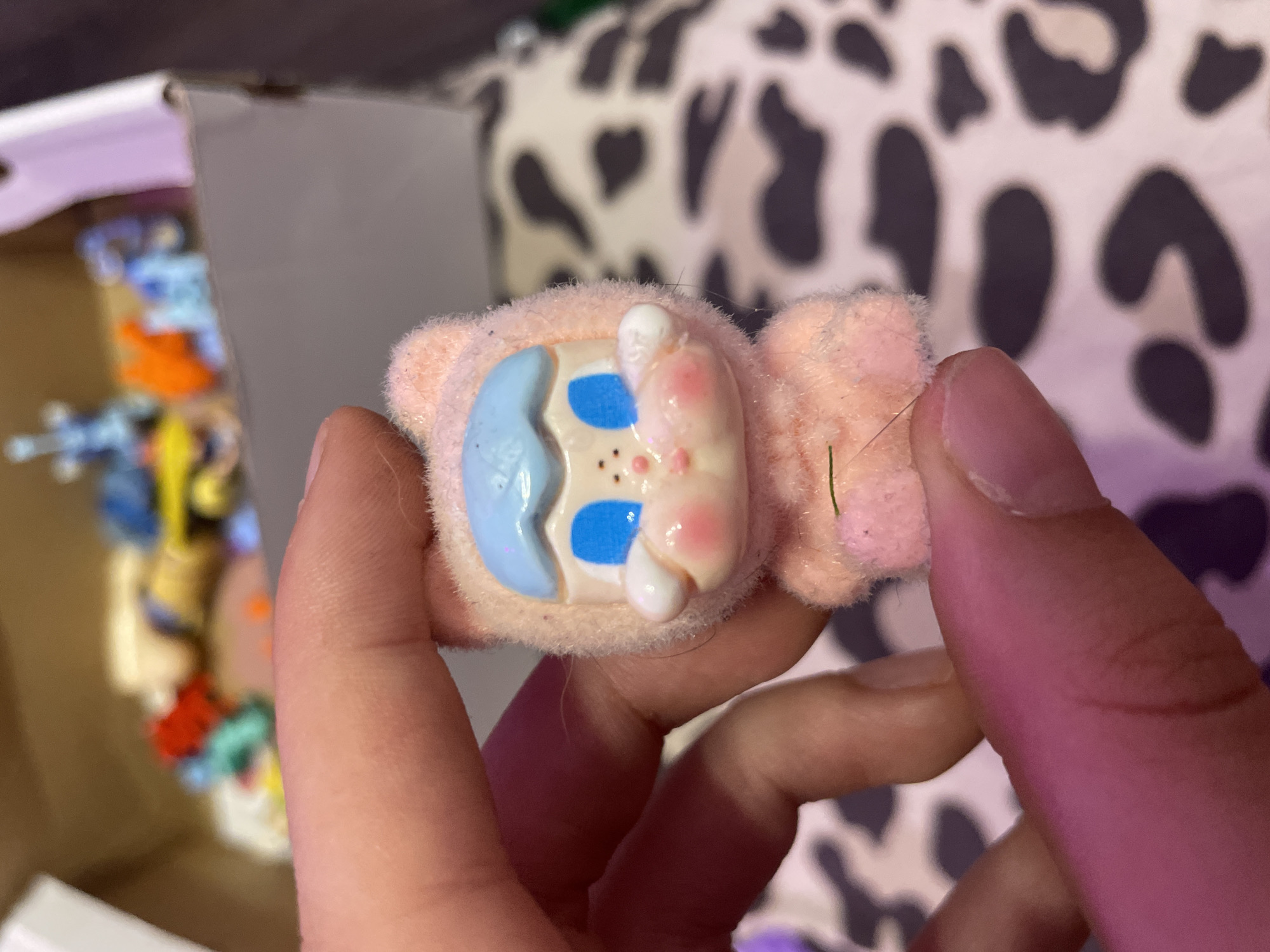 A hand holds a small, fuzzy, pink cat-like figurine with blue eyes and rosy cheeks. The figurine is wearing a light blue hat. In the background, a box contains various blurred colorful miniature toys, including blue, red, and orange figures. The surface behind the figurine appears to be covered with a white and black cow-print fabric. There are no discernible landmarks or context to indicate a location within Ladozhskii Trudposelok, Russia.