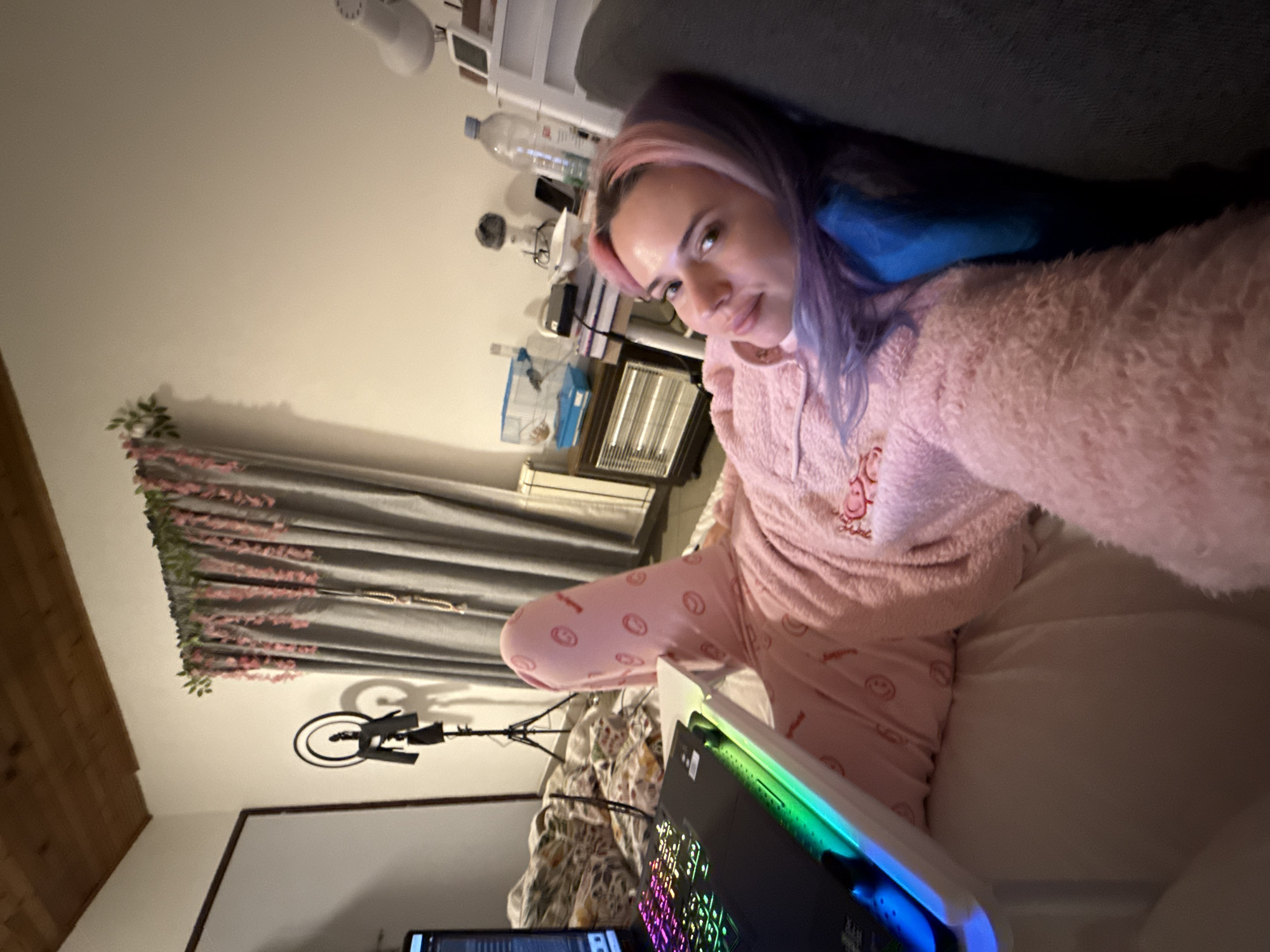 A young adult female is positioned on her back, facing the camera, her head resting against a dark grey cushion. She has light skin, brown eyes, and multi-toned hair with pink and purple hues. She is wearing a pink, fluffy hooded sweatshirt and pink pajama pants adorned with a repeating circular motif. Her left leg is bent at the knee, with her right leg extended. A black laptop with a multi-colored (RGB) backlit keyboard rests on a white, portable stand situated over her legs.
The scene is an indoor, residential room, identified as being in the City of Zagreb, Croatia. To the left of the subject, a patterned duvet covers an unmade bed, behind which a black ring light on a tripod stand is visible. The wall is white, and the ceiling features brown wooden planks. To the right of the subject, a grey curtain hangs, decorated with pink, vine-like floral elements and green leafy accents at the top. Below the curtain, on the floor, is a small, dark brown electric heater. Above the heater, on a small table, sits a blue and white plastic pet cage, a stack of books, a black electronic device, a white bowl, and a black smartphone. Further to the right, a large clear plastic water bottle and a smaller bottle are on a surface next to a white tiered shelf unit, which holds a white desk lamp.