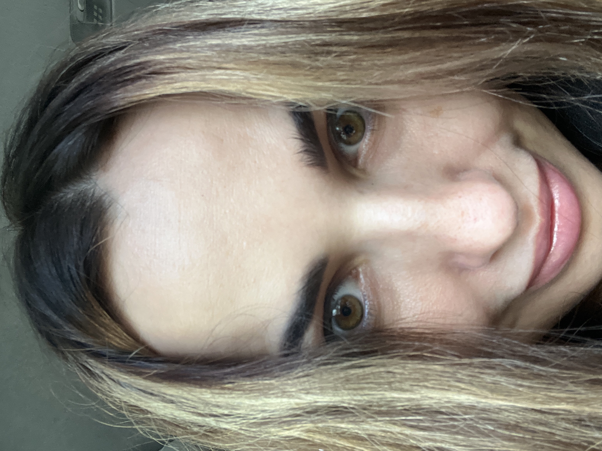 A close-up, rotated portrait shows a person's face, primarily focusing on their eyes, nose, and part of their mouth, framed by hair. The image is oriented with the person's head tilted approximately 90 degrees to the left.
The subject has wide, engaging brown eyes with dark, well-defined eyebrows. Their nose is visible, and the corner of their mouth is turned upwards in a gentle, warm smile, revealing pink, glossy lips. The skin appears smooth with natural texture. A small, light brown mole or freckle is visible on the skin near the right eye's inner corner.
Hair frames the face, showing a blend of dark brown roots transitioning into lighter, warm blonde or light brown highlights, suggesting a balayage or ombré style. The hair appears to be long and falls around the face, with some strands covering parts of the forehead and cheeks. The background is softly out of focus and nondescript, appearing as a plain, light grey surface, with a faint, dark object visible in the upper left corner of the original image, which could be part of an indoor fixture or vehicle interior.
The overall impression is that of a direct, friendly gaze, possibly a selfie. There are no visible objects other than the person and their hair, and no discernible text. Time of day and specific location cues are not apparent.