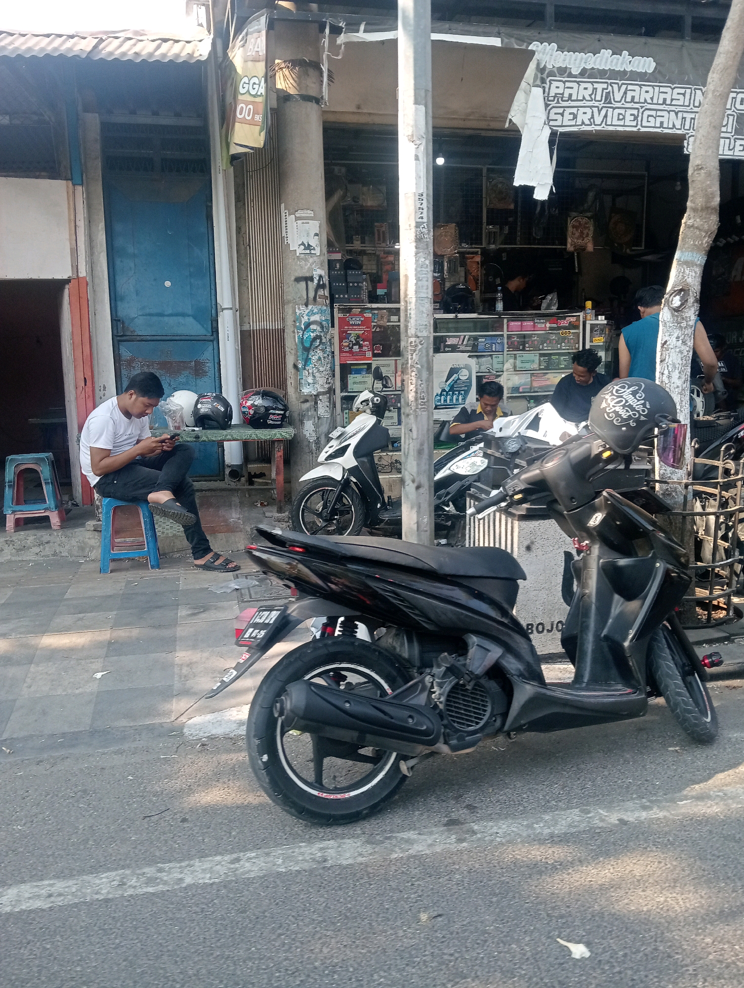 A young man in a white t-shirt and black pants is sitting on a blue plastic stool, looking at his phone. He is wearing black sandals. Next to him on a table are three helmets: a white one, a black one, and a red and black one. In the background, there is a shop with motorcycles and various automotive parts displayed. The shop has a banner that says "Menyediakan PART VARIASI MOTOR SERVICE GANTI...". There are other people inside and outside the shop. Several motorcycles are parked in front of the shop, and one is in the foreground of the image. The scene appears to be a street in the city of Bojonegoro, Indonesia, as indicated by the text on a motorcycle's license plate visible in the foreground. The environment is outdoors, with a sidewalk and a street. The lighting suggests it is daytime.
