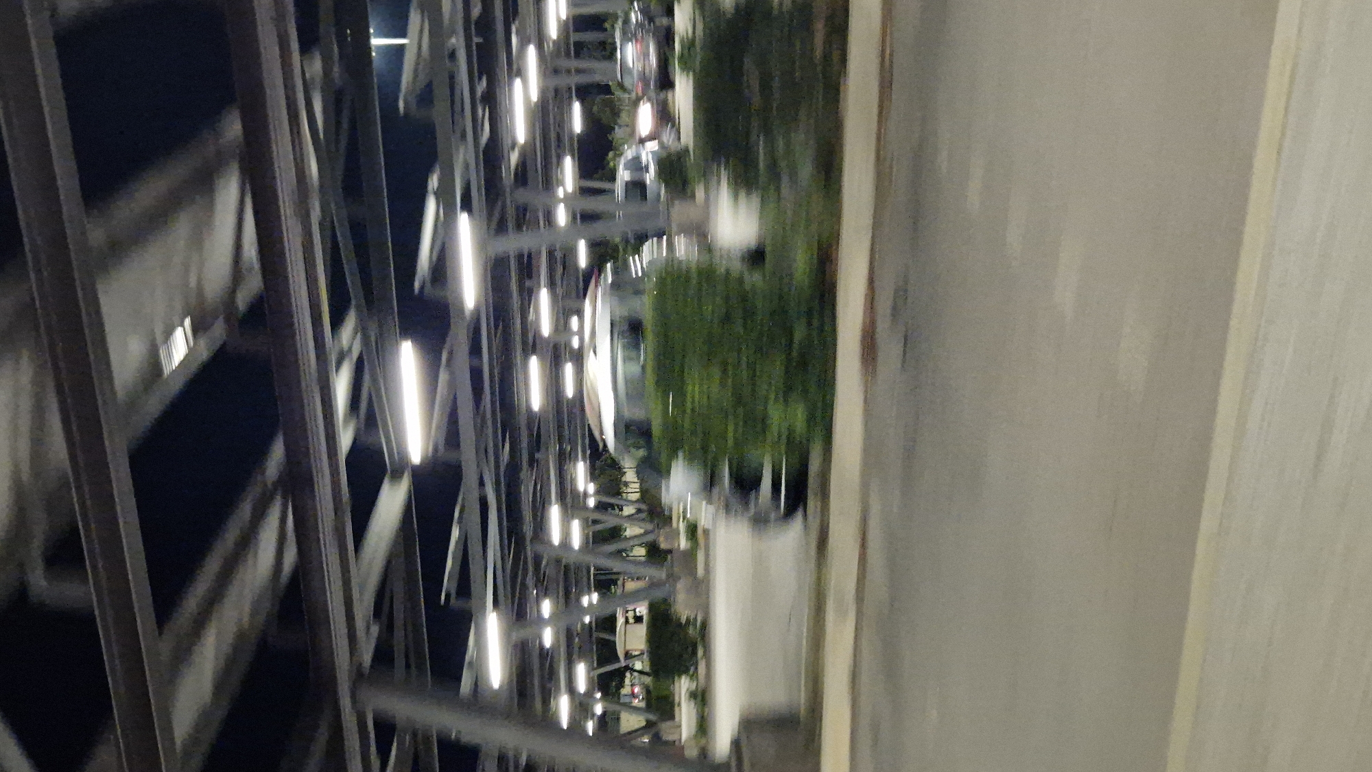 The image captures a blurry nighttime view of a modern, open-air parking structure in Taranto, Italy. Rows of bright, linear lights illuminate the scene from above, casting a stark glow on the metal framework of the structure and the surrounding paved area. The lighting suggests this is likely an airport or a large commercial parking lot, designed to be functional even after dark.
In the mid-ground, a silver minivan is visible, its form blurred due to motion, suggesting it is either moving or the photograph was taken with a slow shutter speed. The minivan is partially obscured by a neatly trimmed green hedge. Behind the minivan, other vehicles are faintly discernible, also affected by motion blur and the low light conditions. To the far left, a concrete pillar supports the overhead structure. The ground is paved with asphalt, with faint white markings visible in the foreground, indicating lanes or parking spaces. The overall atmosphere is utilitarian and functional, typical of such infrastructure.