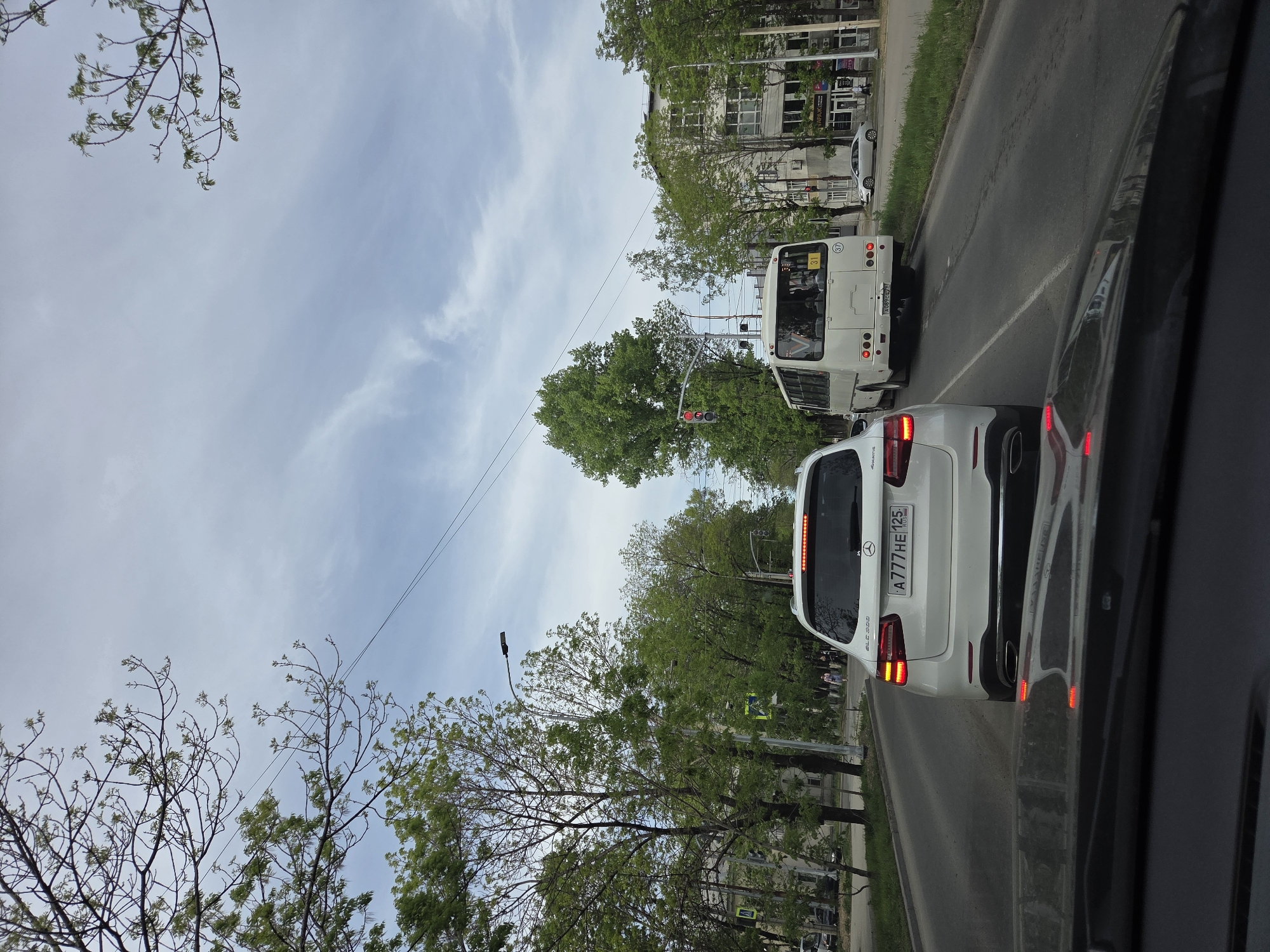 A daytime, street-level view from inside a car, showing traffic stopped at a red light in Birobidzhan, Russia. The car filming the scene is positioned behind a white Mercedes-Benz GLC 300. The Mercedes has the license plate "A777HE125." A white bus is directly ahead of the Mercedes. Buildings and trees line the street. The sky is overcast.