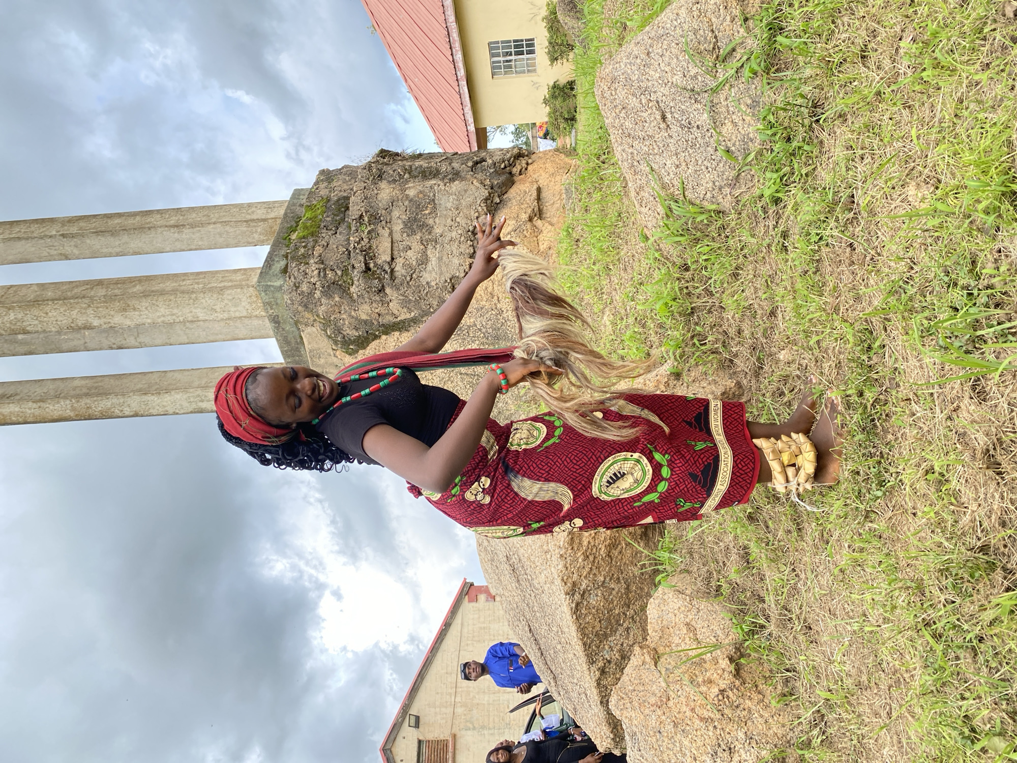 The image shows a young Black woman standing on a grassy hillside next to large rocks, possibly a monument given the concrete pillars in the background. She is wearing a black top, a red headscarf, and a vibrant red skirt with a repeating circular pattern. She has a necklace made of red and green beads. Her hair is braided and black. She is holding a strand of long, blonde and brown hair, possibly a wig or hair extension, in her right hand and smiling broadly. On her right ankle, she has a decoration made of woven yellow material. In the background, a building with a red roof and light yellow walls is visible, along with other people in casual clothing. The sky is overcast and cloudy, suggesting it might be daytime. The setting appears to be outdoors, with a mix of natural rock formations and some man-made structures. There is some text visible on a band around the woman's skirt, but it is too small to read clearly.