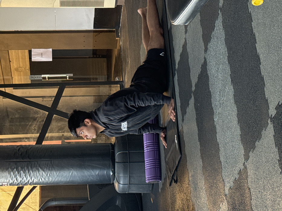 A man in a black hoodie and shorts is on the floor of a gym, performing a forearm plank on a purple foam roller on top of a black exercise mat. The floor is covered in patterned grey and black mats. To the left is a black punching bag. In the background is gym equipment and paneling.