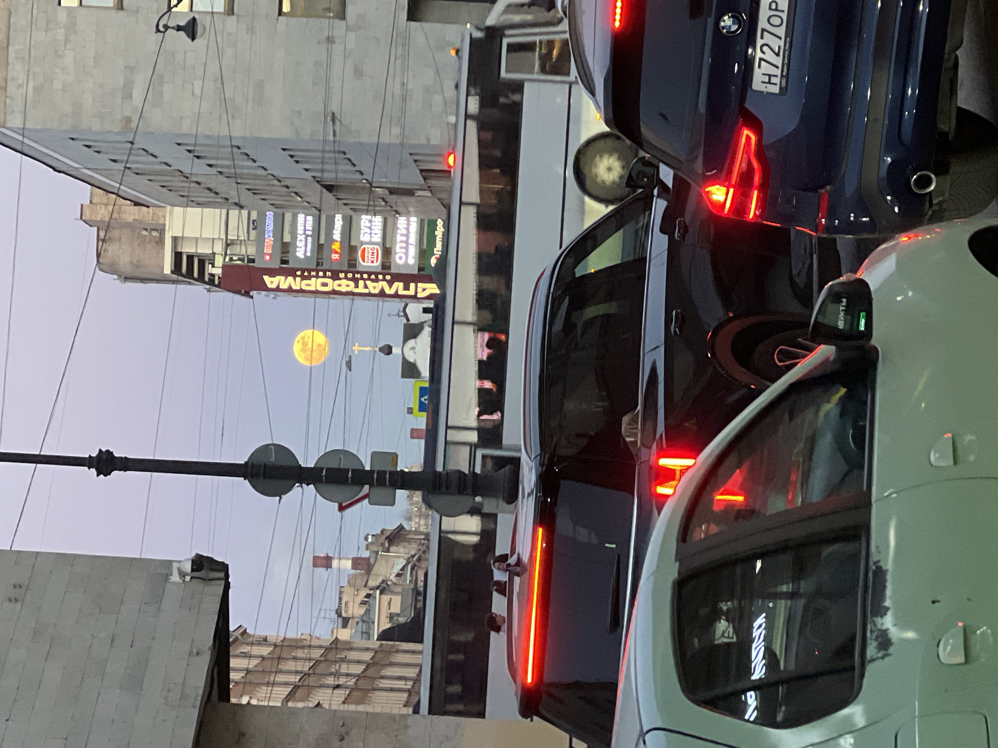 The image is a street scene in Saint Petersburg, Russia, photographed vertically. In the background, there's a building with a prominent sign "ПЛАТФОРМА" (Platform), followed by other signs including "ALEX UTREC", "Burger King," and "ОПТИК" (Optics). In the distance, a yellow moon is visible in the sky. The foreground is occupied by traffic, with multiple cars lined up, including a dark blue BMW with the license plate "H727OP." A bus or large vehicle is visible between the cars, and a light green car is partly visible in the lower right corner. Street signs and power lines are also visible against the sky. The time of day appears to be dusk.