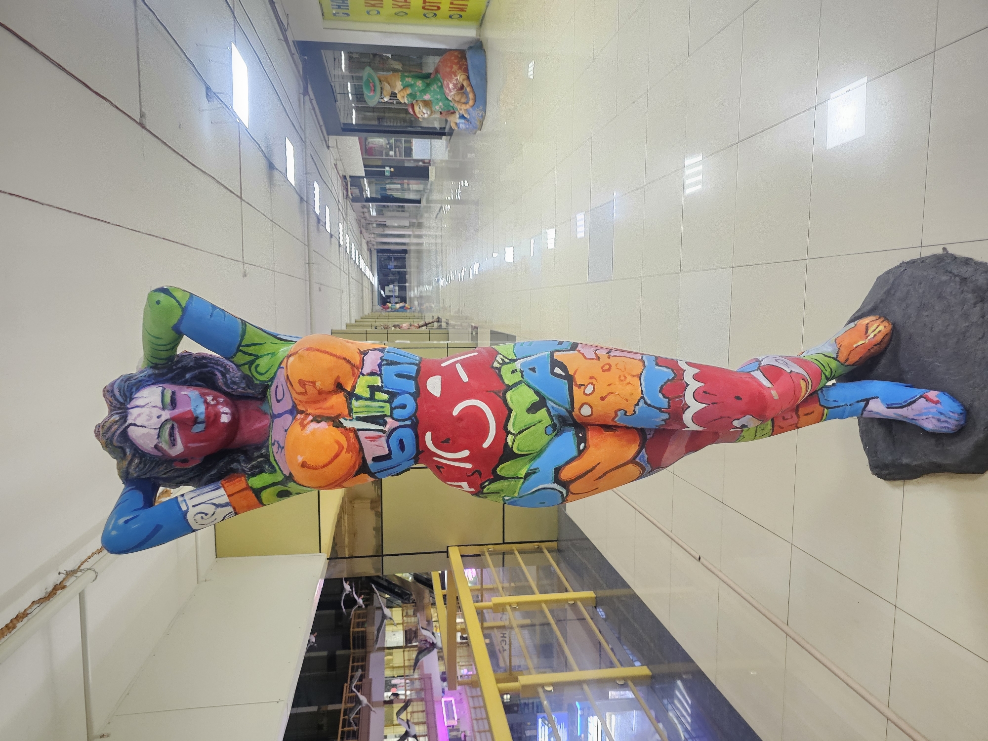 The image shows a colorful graffiti-covered statue of a woman in a shopping mall. The statue is the main subject in the foreground, with its vibrant paint drawing attention. Behind the statue, the mall interior is visible, featuring a long, well-lit corridor lined with storefronts. The floor is tiled, and overhead fluorescent lights illuminate the space. In the distance, there are other decorative elements and storefronts, as well as a second statue depicting two cats. The scene appears to be in an indoor public space.