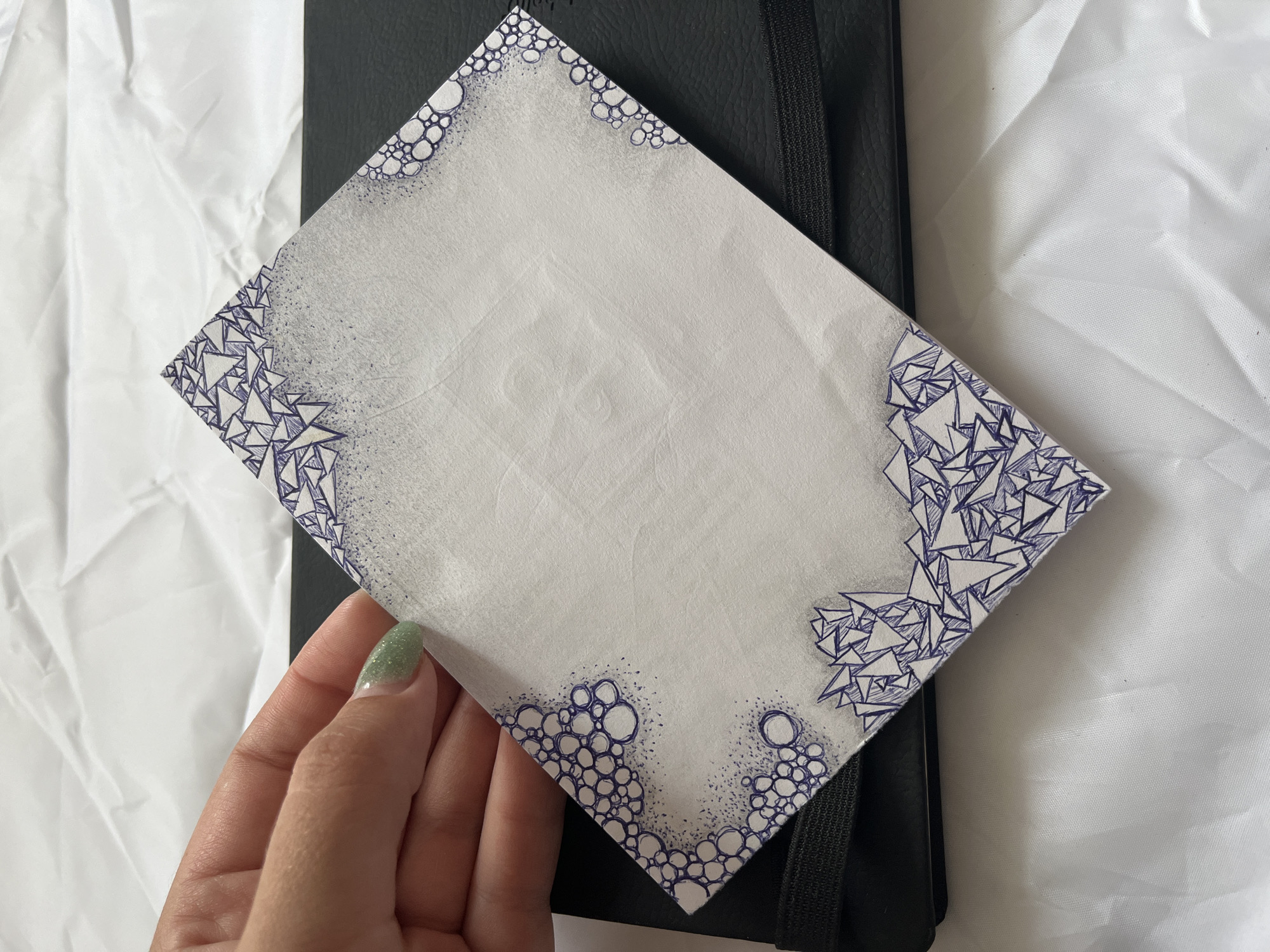 A hand with light green glitter nail polish is holding a rectangular piece of white paper at an angle. The paper has a decorative border of small blue circles and jagged blue triangular shapes. The paper is placed on top of a black book with a black elastic band. The background is a white wrinkled fabric. There is no indication of the location in Krasnyye Barrikady, Russia.
