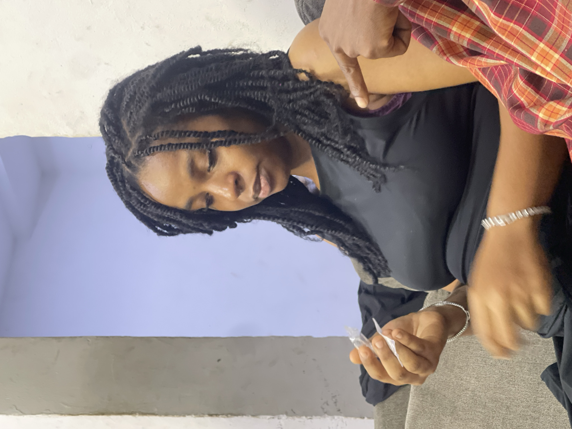 An overhead, medium shot shows a young Black woman with long, dark, twisted braids sitting on a gray couch. She is wearing a black tank top and silver bracelets on both wrists. The woman is looking down at a small, clear object she holds in her left hand. A person's right hand, wearing a red and orange plaid shirt, is visible pointing at the woman's shoulder. The background consists of a light blue wall with a white column on the left. The image appears to have been taken indoors, in an urban setting such as Abuja, Nigeria.