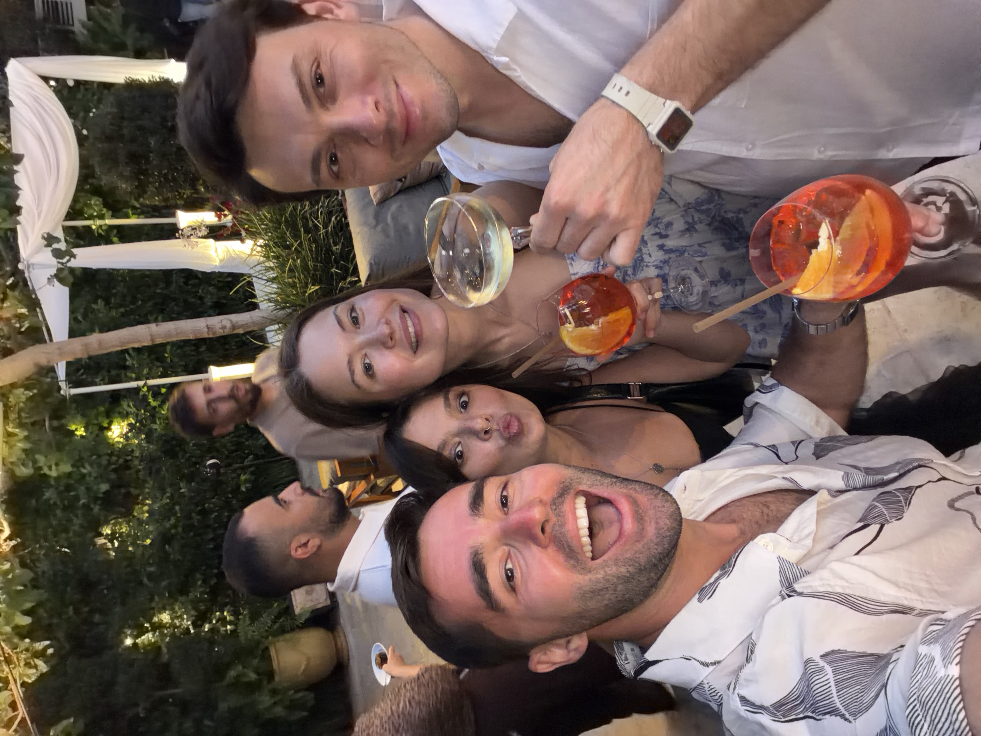 A group of six people is gathered outdoors. Four individuals are positioned in the foreground, with two holding drinks, including orange-colored beverages and white wine, for the apparent selfie photo. Behind them, two additional people are visible in the background, near greenery. The setting includes a decorated tent and foliage, suggesting an outdoor venue, likely in Tel-Aviv, Israel.