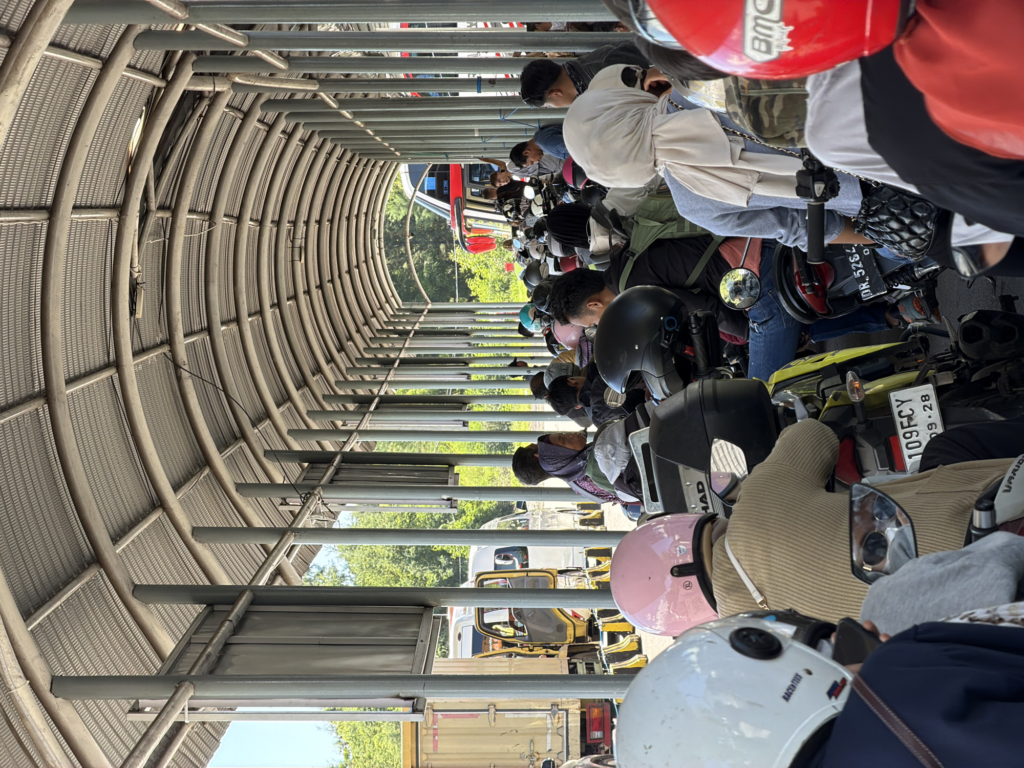 The image depicts a motorcycle parking area, likely a covered tunnel, located in Gunung Malang, Indonesia. A line of motorcycles is parked closely together, each with a rider or passengers. Many of the riders are wearing helmets in various colors, including red, pink, white, and black. The motorcycles have license plates, one of which reads "109FCY". Riders are observed seated on their motorbikes. The tunnel structure is made of metal ribs covered with mesh, and beyond the parking area, trees and glimpses of vehicles, including what appears to be a bus, are visible. The background includes some blurred details of the urban environment.