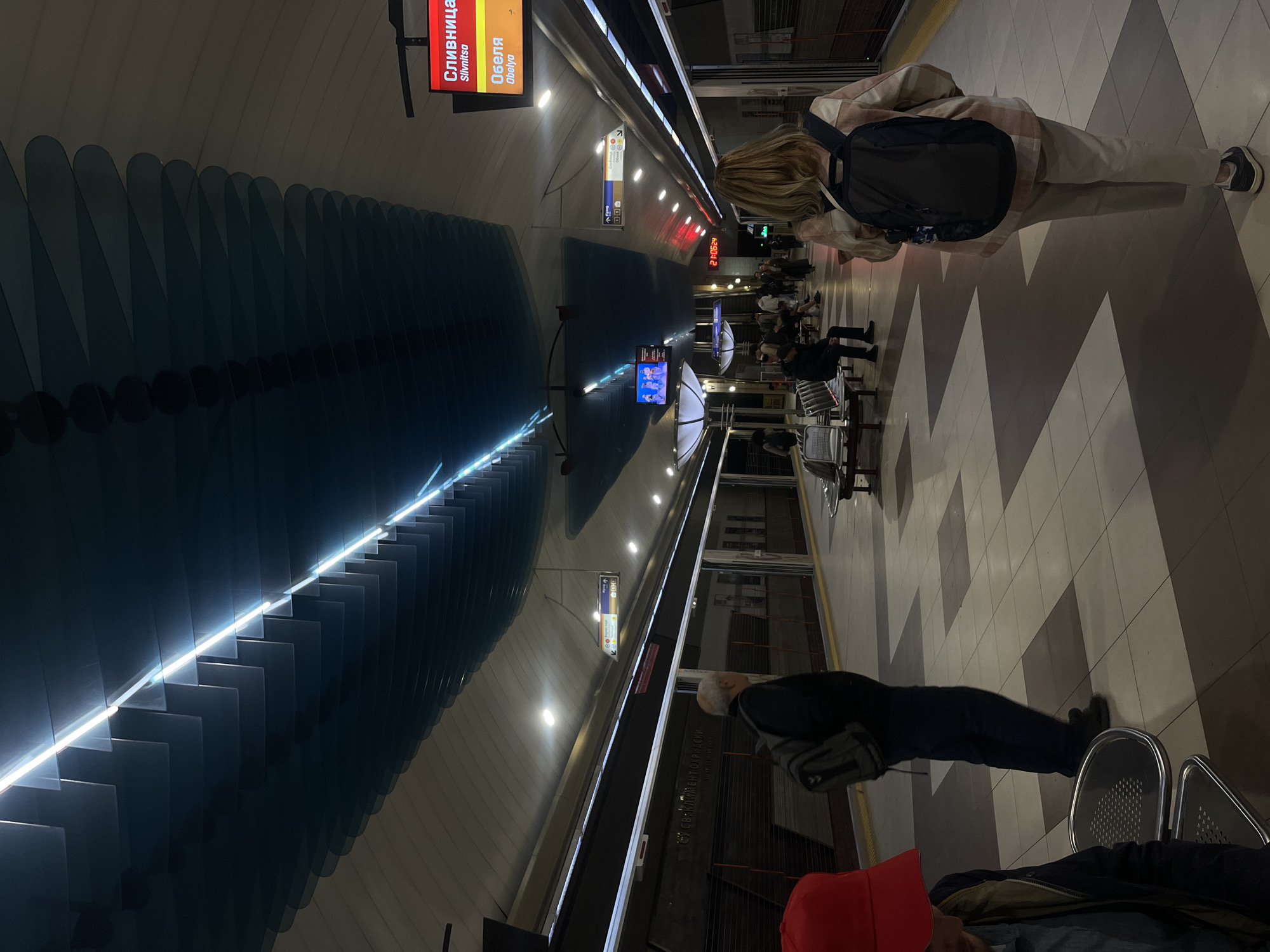 The image shows the Slivnitsa metro station in Sofia, Bulgaria. Several people are waiting on the platform. There are benches for seating, and a large sign indicates the station name and directions. The station features a unique design with layered blue elements on the ceiling, illuminated by linear lights. A display shows the time as 21:35:54. The floor has a geometric pattern.