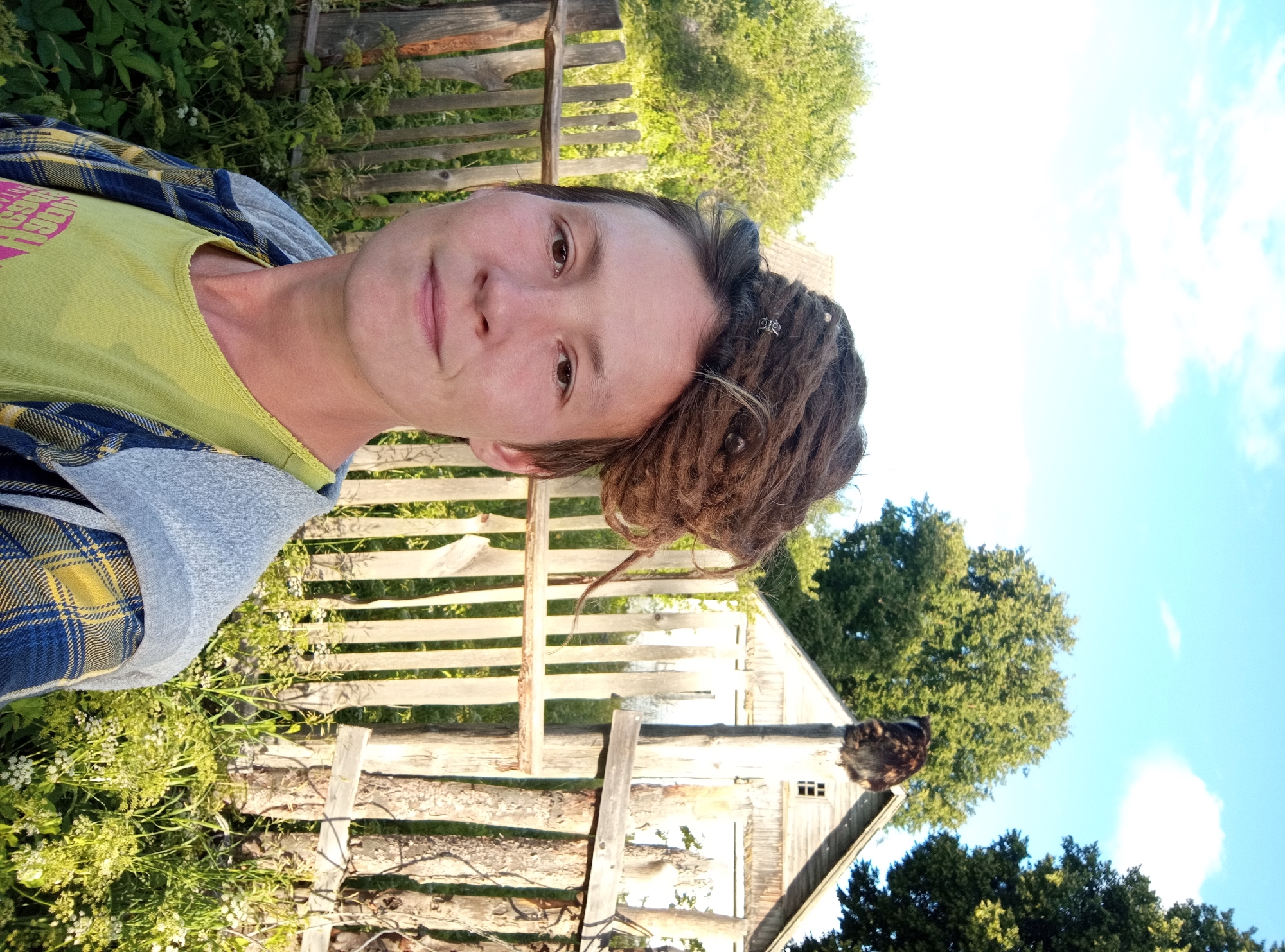 In Ostrovtsy, Russia, a woman with dreadlocks styled into a bun on top of her head stands in front of a wooden fence and a rustic building. She is wearing a lime green t-shirt and a blue and yellow plaid jacket. Behind her, a cat is perched on top of a wooden post. The scene is set outdoors with green trees and a blue sky with clouds.