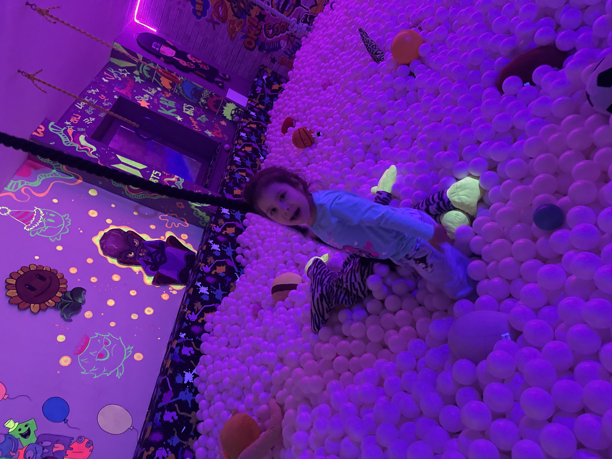 An indoor play area features a young girl submerged up to her waist in a large ball pit. The pit is filled with countless white plastic balls, which appear vibrant purple under the dominant blacklight illumination. The girl, wearing a light blue long-sleeved top and light pants, is seated, facing the camera with a slight smile and open mouth. Various toys are scattered within the balls, including an orange octopus-like plush, a striped animal plush with neon green feet, a basketball, and other colorful objects.
The walls surrounding the ball pit are dark purple, adorned with extensive glow-in-the-dark artwork. The left wall displays outlines of cartoon characters, including a sunflower and an angry green creature resembling characters from "Plants vs. Zombies," along with balloons and glowing yellow dots. The right wall is covered in graffiti-style art, featuring K-Pop band names such as "BTS," "Stray Kids," and "Enhypen," abstract designs, and other glowing text in neon green, yellow, and orange. A pink neon light strip runs along the upper right wall. Two sets of ropes with wooden handles and one ring, part of a suspended play structure, hang from the ceiling in the mid-ground. The overall lighting is dim, primarily provided by UV blacklights, which cause the balls and painted wall elements to fluoresce brightly.
Location: Unknown, Unknown.