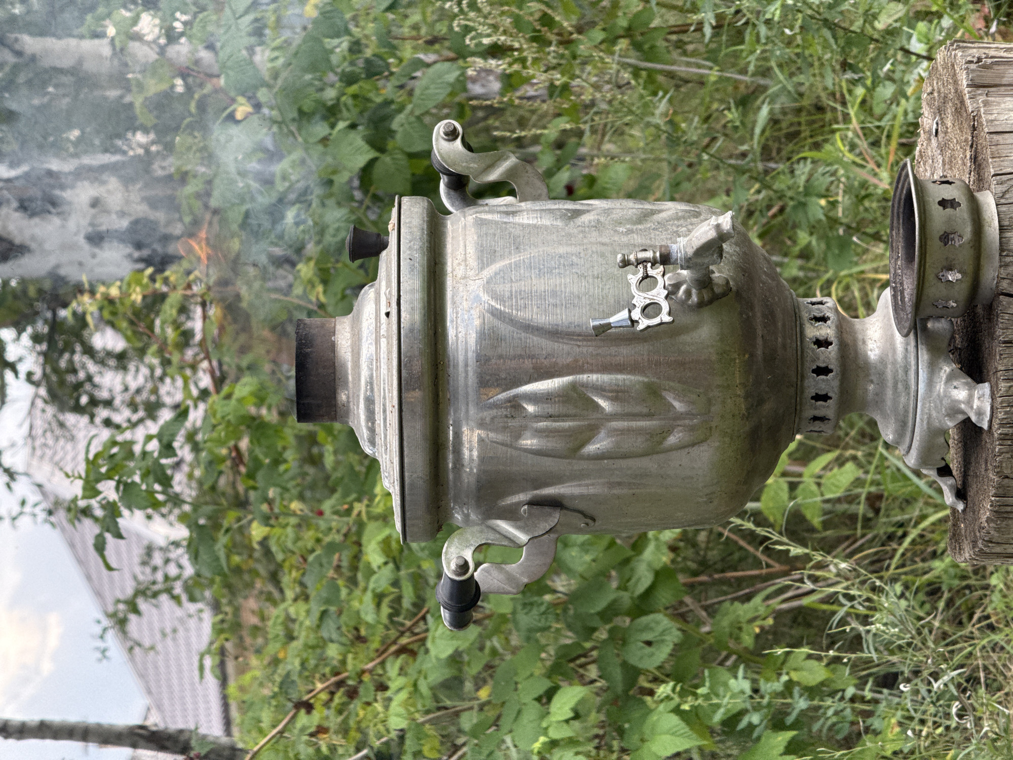 An outdoor scene features a traditional Russian samovar made of metal, positioned on a weathered wooden stump. The samovar is actively in use, evidenced by smoke and a small flame visible from its top. It has decorative leaf patterns on its body and a spigot for dispensing hot water. Below the samovar, there is a separate metal container, likely for fuel. The background is a dense growth of green foliage, suggesting a garden or natural setting. In the upper left corner of the background, a portion of a roof is visible, indicating the presence of a dwelling. The overall impression is one of a rural or semi-rural environment, potentially in or around Kremenkul', Russia, given the context. There are no people or animals present in the image.