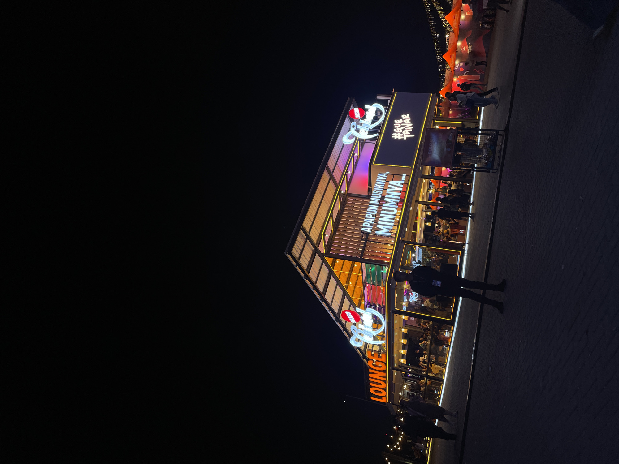 At night in Salembaran, Indonesia, a brightly lit establishment, labeled "LOUNGE" with an upper-level sign reading "APAPUN MUSIKNYA, MINUMNYA..." and "#GUETANAK", stands out against the dark sky. The building has an open, multi-level design, with patrons visible inside through glass walls. A person stands on the sidewalk outside, their shadow stretching onto the pavement. In the upper right, a partially visible, tent-like structure is illuminated. The environment is dark with a paved surface extending from the building.