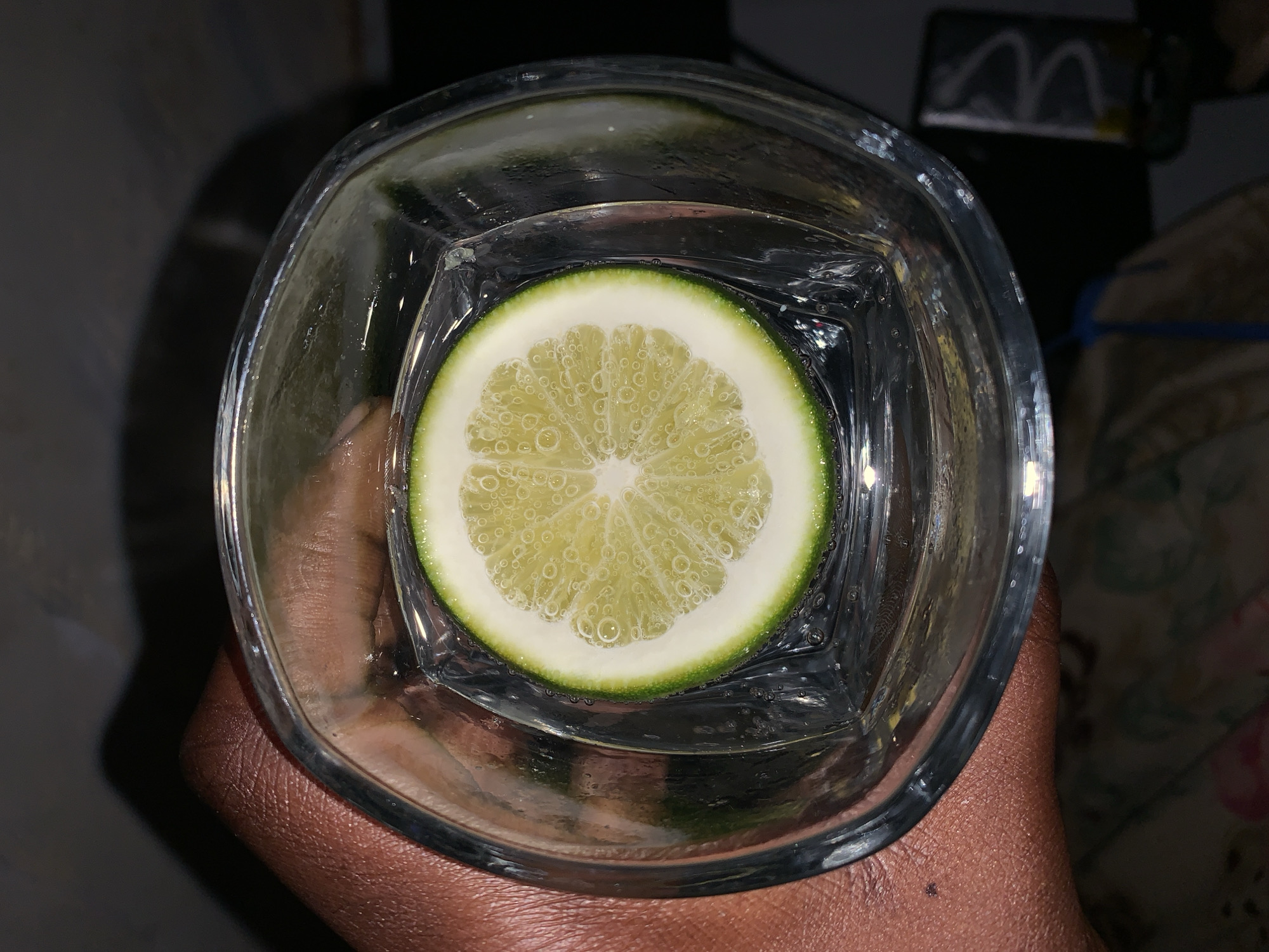 A person with dark skin is holding a square glass containing a clear liquid with a lemon slice in it. The lemon slice is round with a green rind and a light yellow interior with segments radiating from the center. Small bubbles cling to the lemon slice. The glass is partially filled with the liquid, and the top of the liquid line is visible in the upper part of the glass. The background includes a blurred dark surface and a hint of an object with a recognizable "M" logo. The location information "Rafin Par, Nigeria" appears to be incorrect and not related to the visual content of the image.