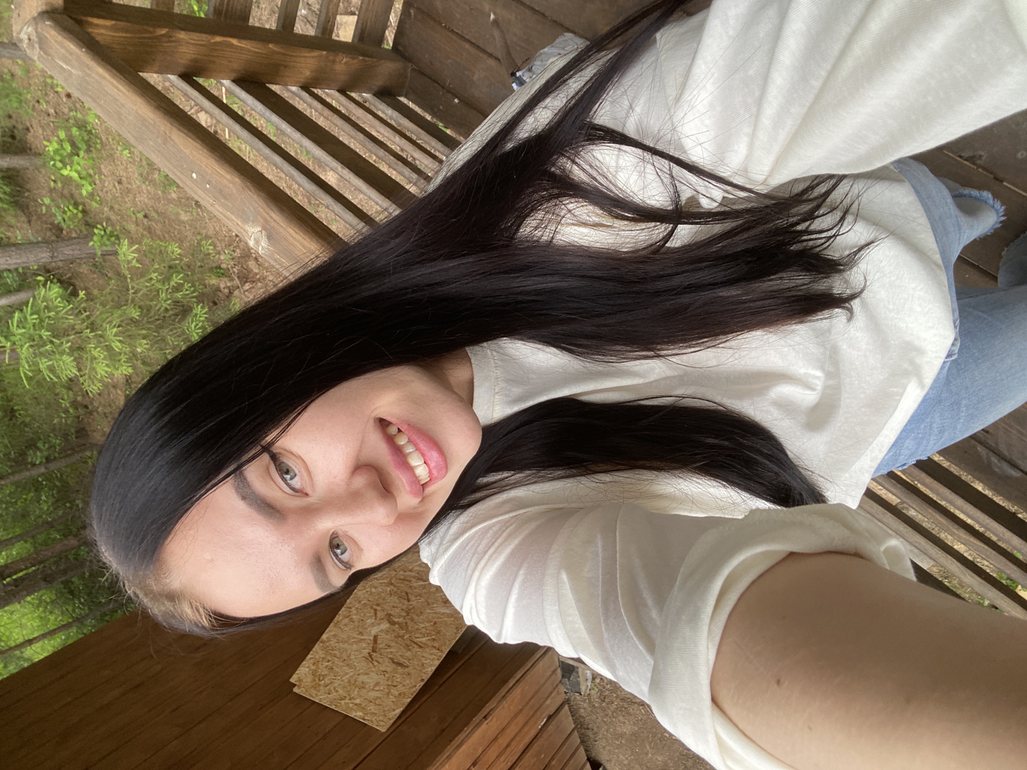 A young woman with long, dark hair and green eyes smiles at the camera. She is wearing a white long-sleeved shirt and blue jeans. She is standing on a wooden deck with a railing, and a forest can be seen in the background. The image appears to be a selfie taken in a natural outdoor setting, possibly in the city of Fokino, Russia, given the presence of evergreen trees common in that region. The lighting suggests it is daytime.