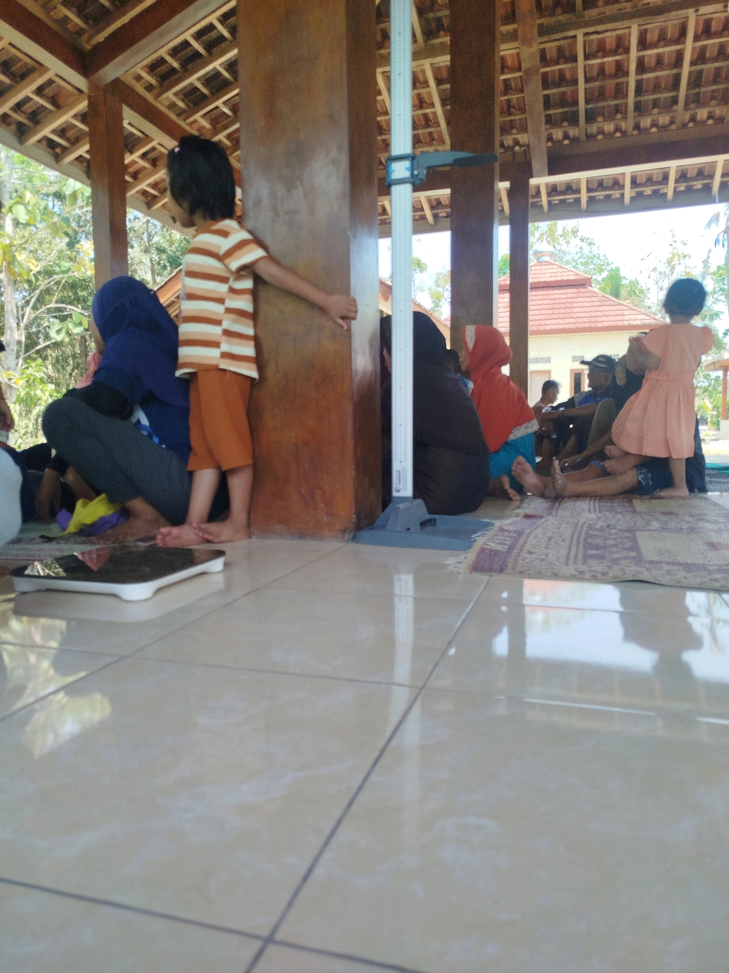 In Semin, Indonesia, a group of people, including children, are gathered under a wooden-roofed structure with a tiled floor. A young child, wearing a striped shirt and orange shorts, stands next to a stadiometer, suggesting a health check or measurement is taking place. Several other individuals are seated on a rug in the background, some appearing to be adults. The scene is outdoors, with trees and buildings visible in the distance. A digital scale is placed on the tiled floor in the foreground. The overall atmosphere appears casual and community-oriented.