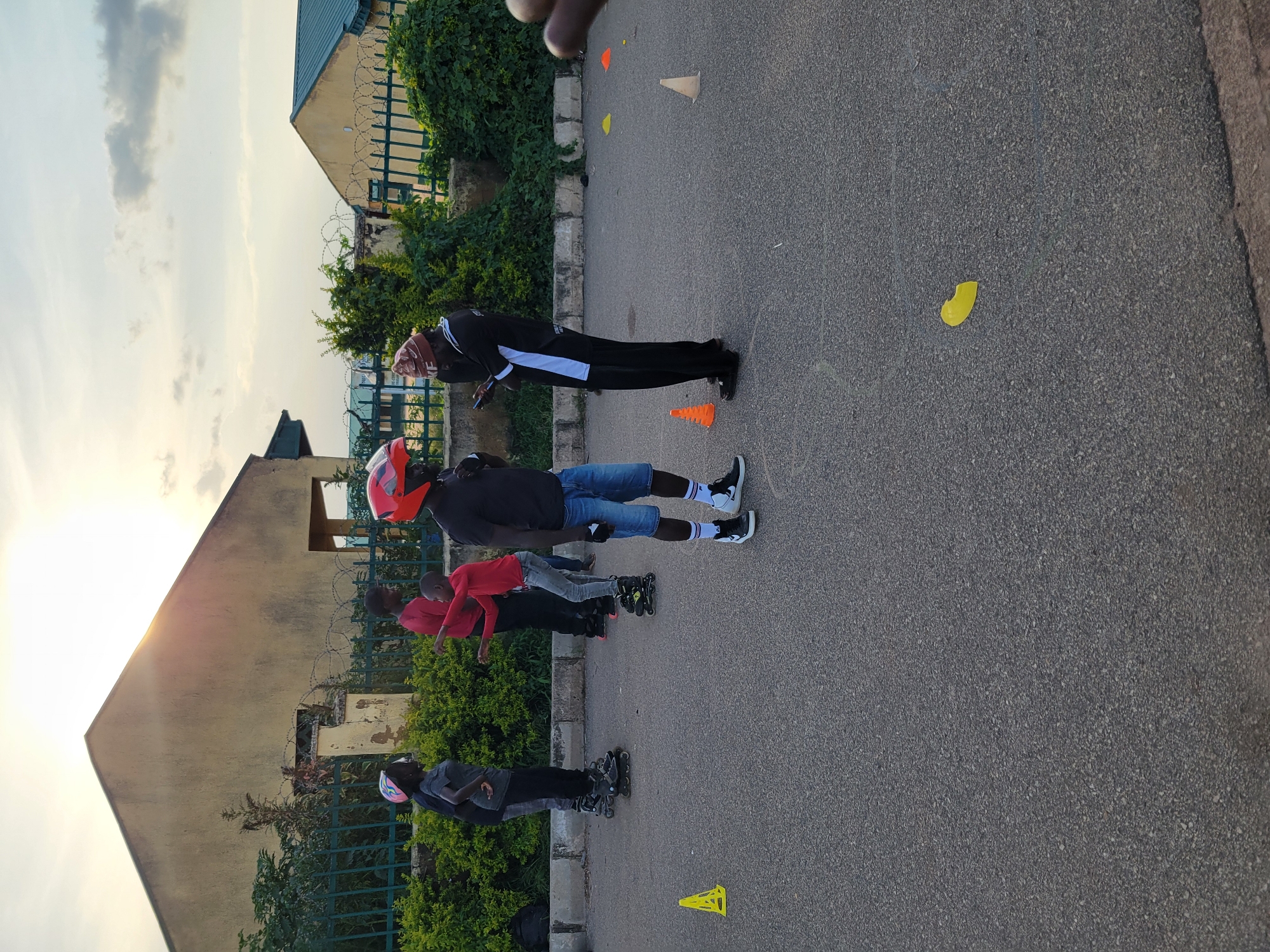In an outdoor setting on asphalt, likely in Jos, Nigeria, a group of individuals are engaged in inline skating. The scene captures a late afternoon or early evening light, with the sun casting a warm glow in the sky.
In the foreground, a person wearing an orange helmet, a black t-shirt, and denim shorts is standing on inline skates. To their left, a younger individual in a red long-sleeved shirt and gray pants is also on skates, with their arms outstretched as if in motion or interacting with someone off-camera. Further left, two individuals are visible; one in a gray t-shirt and black pants, and another behind them, partially obscured.
In the background, a building with a yellow facade and a green roof stands, partially covered by lush green foliage. A fence made of green metal bars runs in front of the vegetation. Scattered on the asphalt are several traffic cones in yellow and orange, and a single yellow leaf. The overall atmosphere suggests a recreational activity taking place in an urban or semi-urban environment.