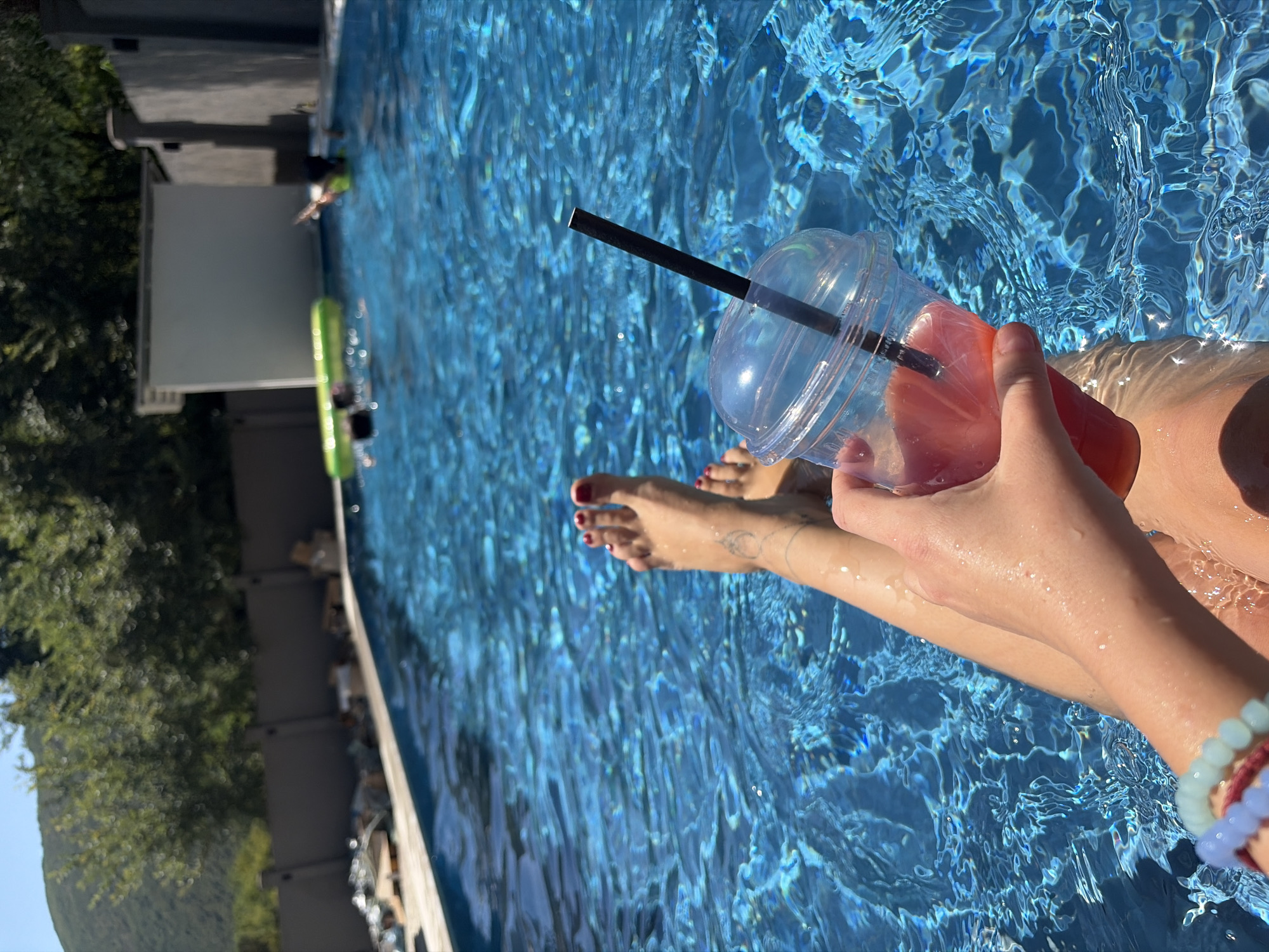 A person's legs and feet are submerged in a bright blue swimming pool. The person is holding a clear plastic cup with a red-colored drink and a black straw. The water is rippling and reflecting the sunlight. In the background, there is a gray fence, trees, and a white building. Two other people are visible in the pool, one on a green inflatable tube and another swimming. The location is Lastva Grbaljska, Montenegro.