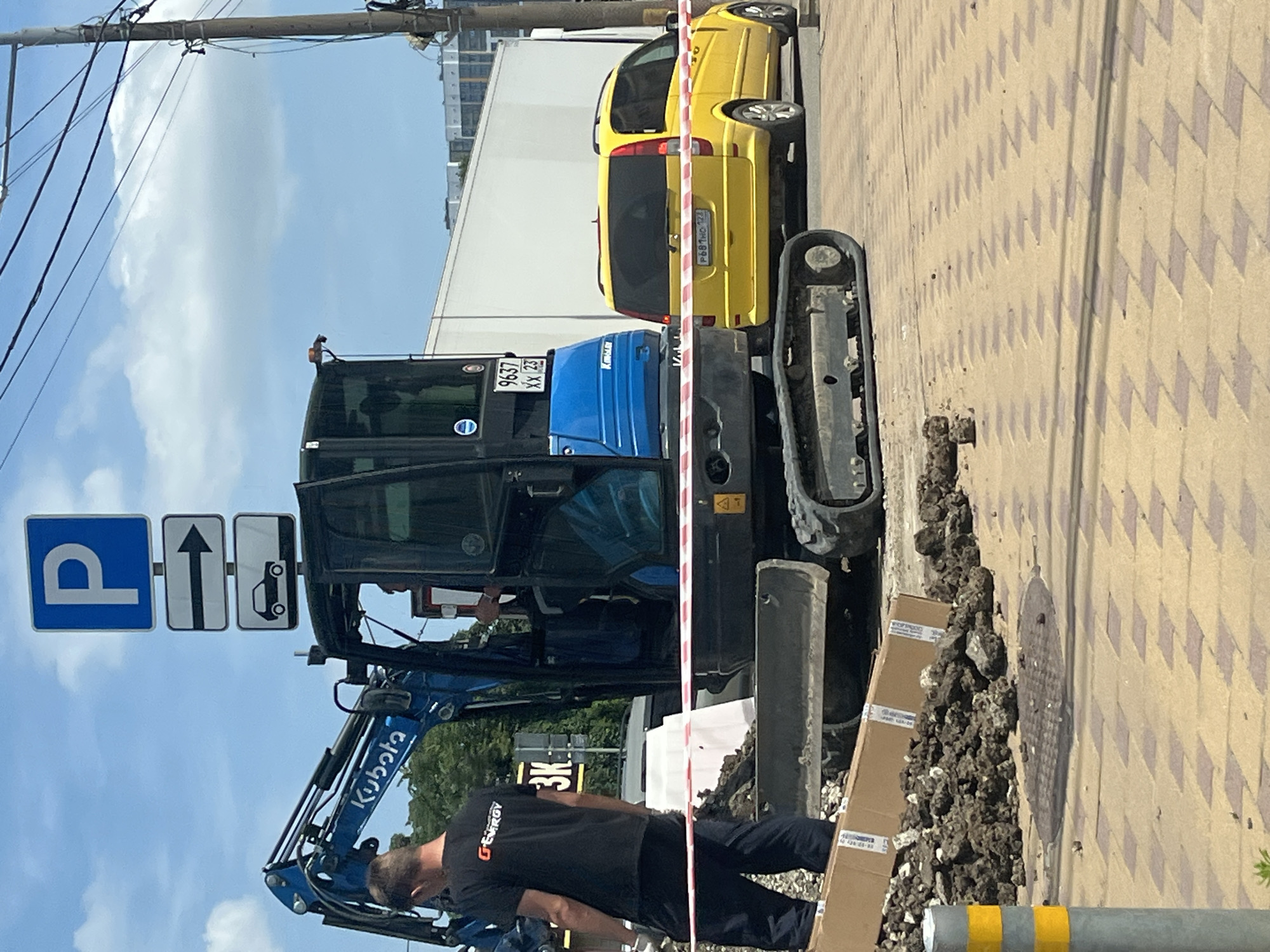 In the city of Krasnodar, Russia, on a sunny day, a mini excavator is parked on a paved area, possibly a sidewalk or road under construction. A person wearing a black t-shirt with an orange logo that says "G-ENERGY" and dark pants is standing to the left of the excavator, facing away from the camera. The excavator is blue and has the brand name "Kubota" visible on its arm. A license plate on the excavator reads "9637 XX 23". A yellow minivan is parked behind the excavator. Traffic signs are visible in the background, including a parking sign and an arrow pointing to the right. Red and white caution tape stretches across the scene, indicating a restricted area. Piles of dirt and debris are scattered around the excavator, and a manhole cover is visible on the ground. A yellow and grey pole is on the left side of the image. The environment appears to be urban, with buildings visible in the distance and power lines overhead.