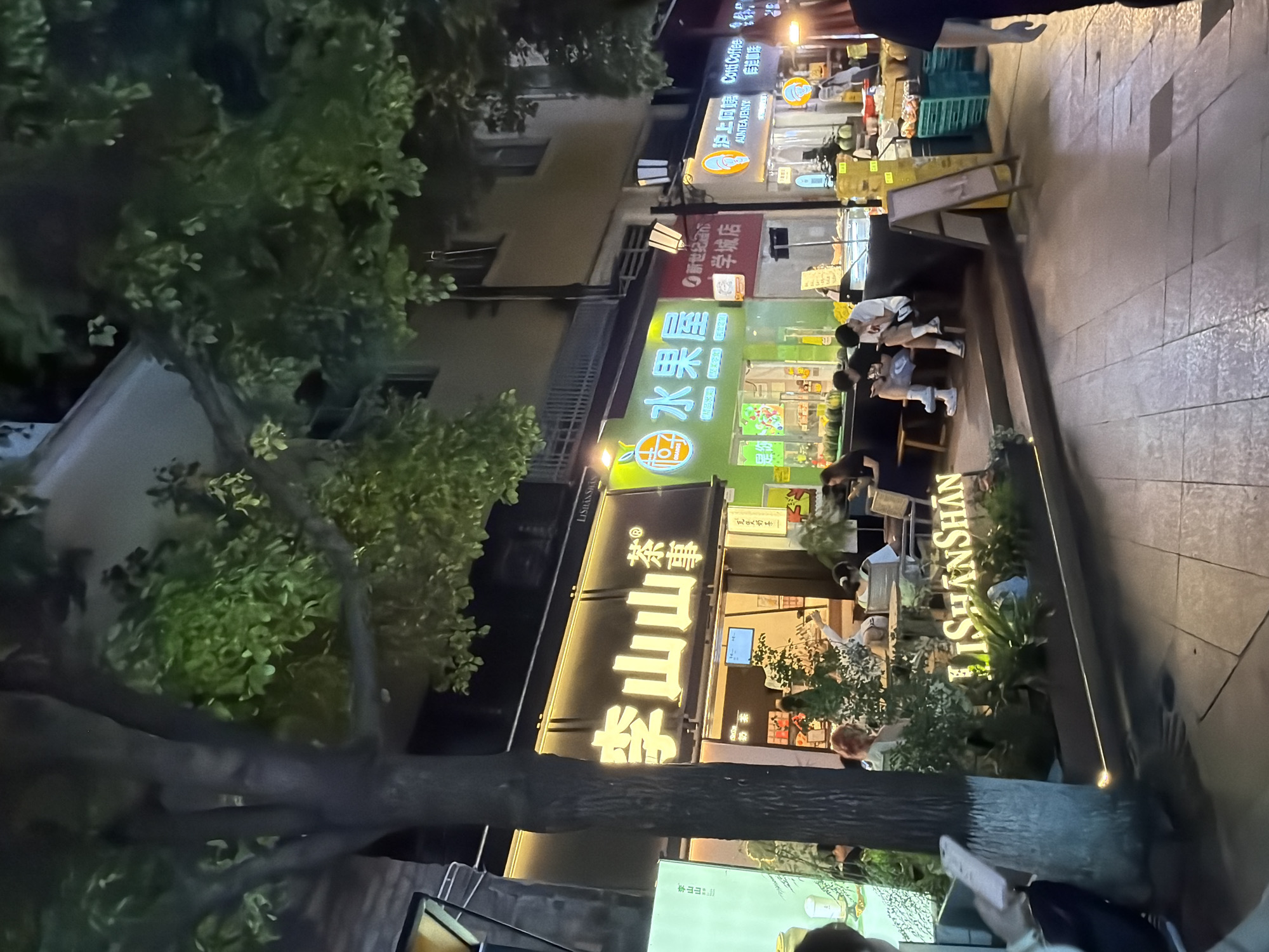 A nighttime outdoor scene shows a street in Chongqing High-tech Zone, China, with several illuminated storefronts and trees. In the foreground, a person is partially visible on the left, holding a phone. To the right, a neon sign displays "LISHANSHAN" in white capital letters. Behind the sign, a tree trunk is prominent. Several people are seated on benches and at tables outside the businesses.
The storefronts are varied. The leftmost business has a green facade with Chinese characters and illuminated graphics of fruit. Next to it, a shop displays "水果屋" (Shuǐguǒ Wū - Fruit House) in large, illuminated characters on a green background, also featuring fruit graphics. Further to the right, a shop has a sign with Chinese characters and "AUNTIE JENNY" on a lighter background, with an illuminated circular logo. Another sign reads "Cotti Coffee". Green plastic crates are stacked near the shops, likely containing produce. The street is paved with textured tiles. The overall atmosphere suggests a lively commercial street at night.