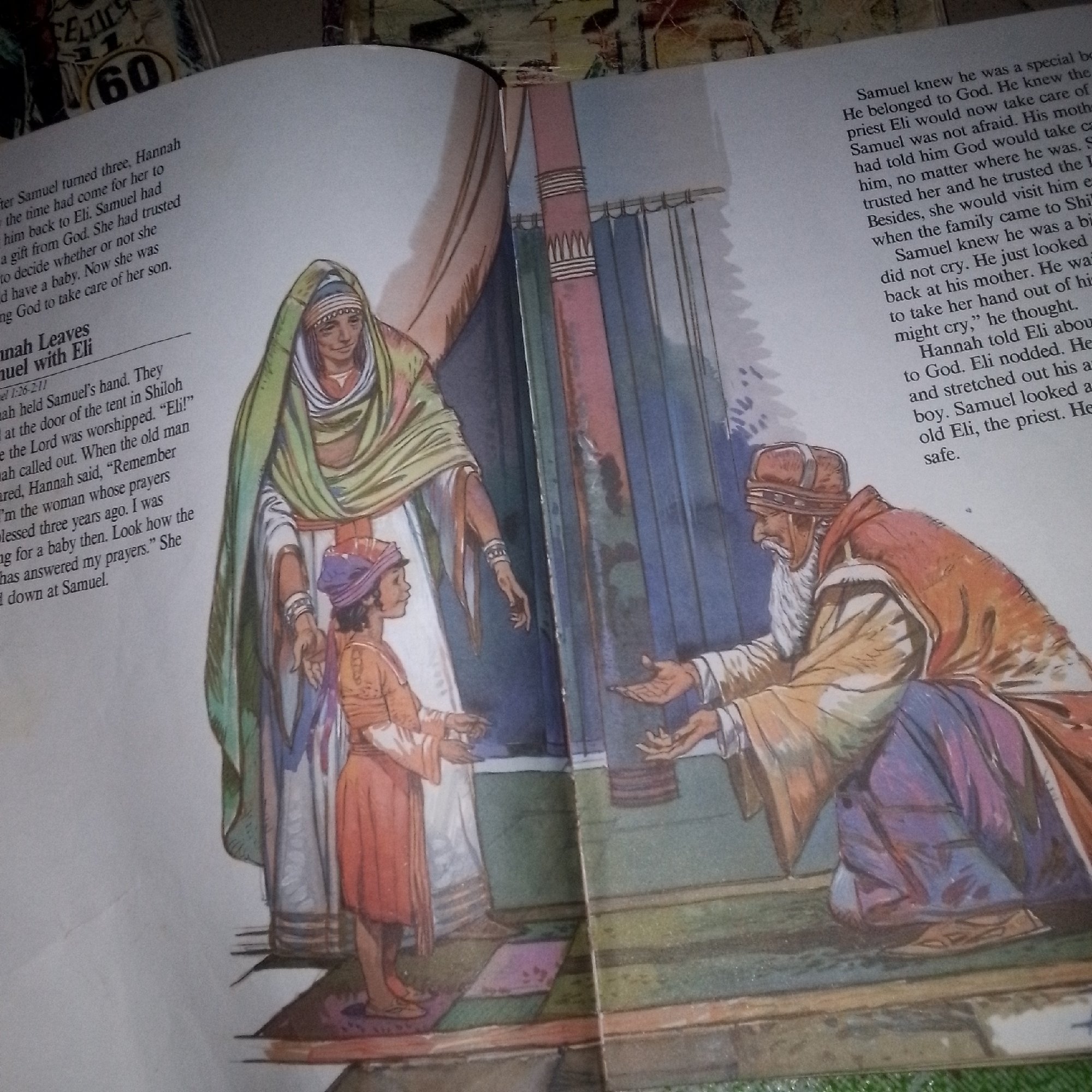 The image is a photograph of an open book with an illustration depicting a scene from the Bible. The illustration shows three people: an older woman in a green and yellow robe, a young boy in an orange tunic and purple head covering, and an elderly man in a brown and orange robe. The woman and boy are on the left side of the illustration, and the elderly man is on the right. The boy is looking at the woman, and the woman is looking down at the boy. The elderly man is reaching out his hands towards the boy and woman. The background consists of colorful curtains and what appears to be part of a tent. Text is visible on both pages of the open book, with headings such as "Hannah Leaves Samuel with Eli" and "Samuel 1:26-2:11". The book is lying on a surface that appears to be a table covered with a fabric. There are no discernible details indicating a specific location in a city.