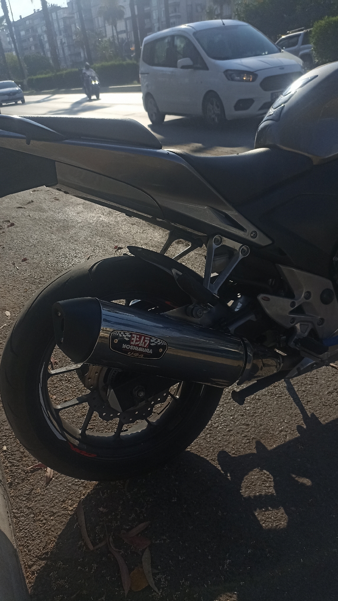 A close-up, low-angle shot focuses on the rear of a dark motorcycle parked on an asphalt surface. The motorcycle's exhaust pipe, prominently featuring a Yoshimura USA logo, is visible. The rear tire and part of the wheel are also in view. In the background, a street scene unfolds under bright sunlight, suggesting daytime. A white van and a car are driving on the road. A person on a motorcycle can be seen further down the road, appearing as a silhouette. Tall buildings and palm trees line the street, indicating an urban or suburban setting. The lighting creates strong shadows on the ground, and fallen leaves are scattered on the asphalt, hints of an autumn or warmer season. The overall mood is casual and everyday, capturing a moment in a street environment. The visible text on the exhaust pipe reads "ヨシムラ" (Yoshimura) and "YOSHIMURA USA".
