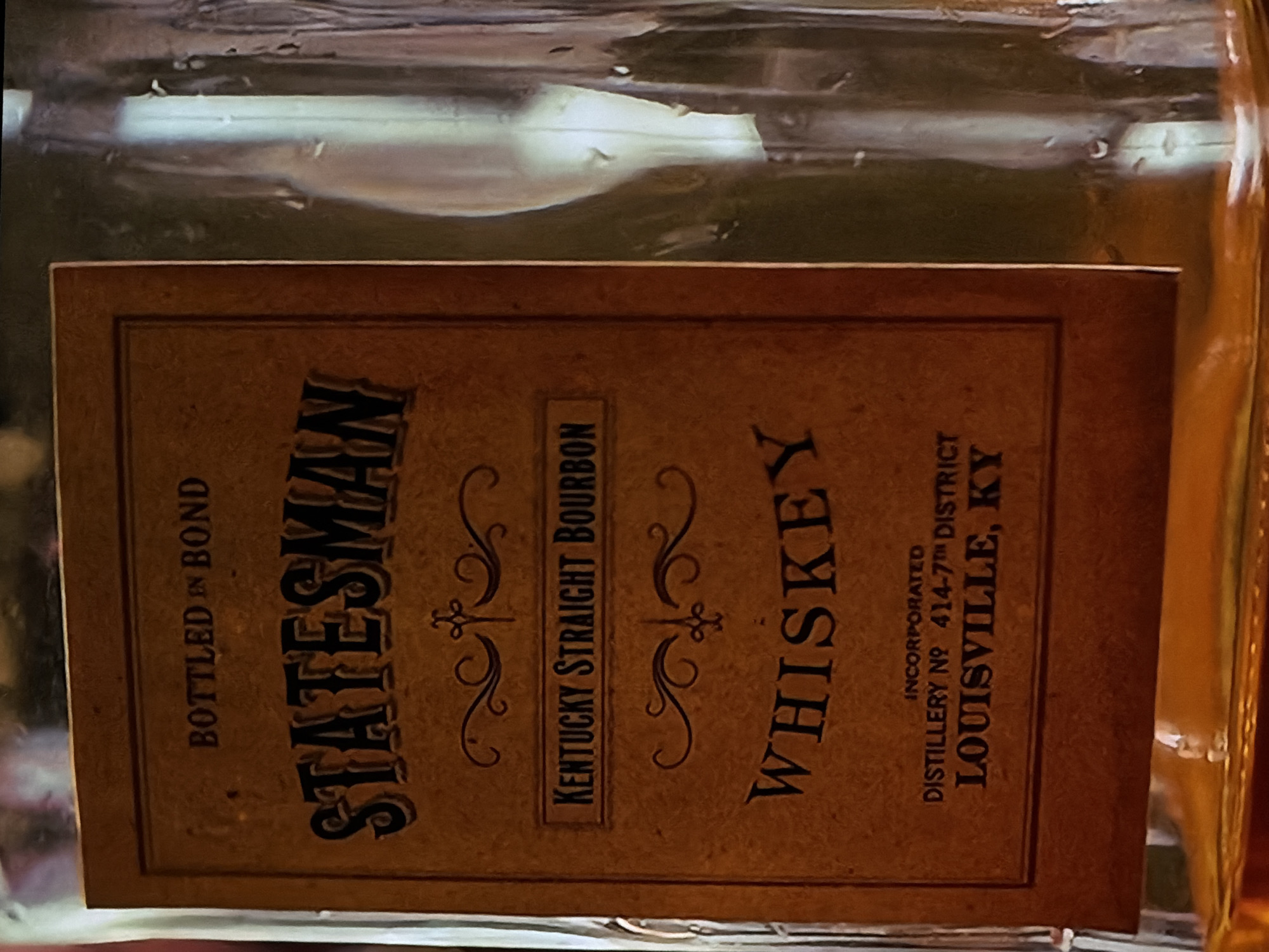 The image presents a close-up, slightly tilted view of a whiskey bottle's label. The label is a rectangular, textured brown or tan paper with dark brown lettering and decorative scrollwork, imparting a vintage aesthetic. Above the label, the clear glass of the bottle is visible, showing reflections, slight distortions, and what appears to be the amber color of the liquid inside.
The most prominent text on the label reads "STATESMAN" in a stylized, bold font. Below this, it specifies "KENTUCKY STRAIGHT BOURBON" and "WHISKEY." Other text includes "BOTTLED IN BOND" along the left side. On the right side of the label, smaller text indicates "INCORPORATED" and "DISTILLERY Nº 414-7TH DISTRICT LOUISVILLE, KY," identifying the origin and specifics of the product. No activity is taking place; the image is a static shot of the bottle. The photograph was captured in Sofiivska Borshchahivka, Ukraine.
