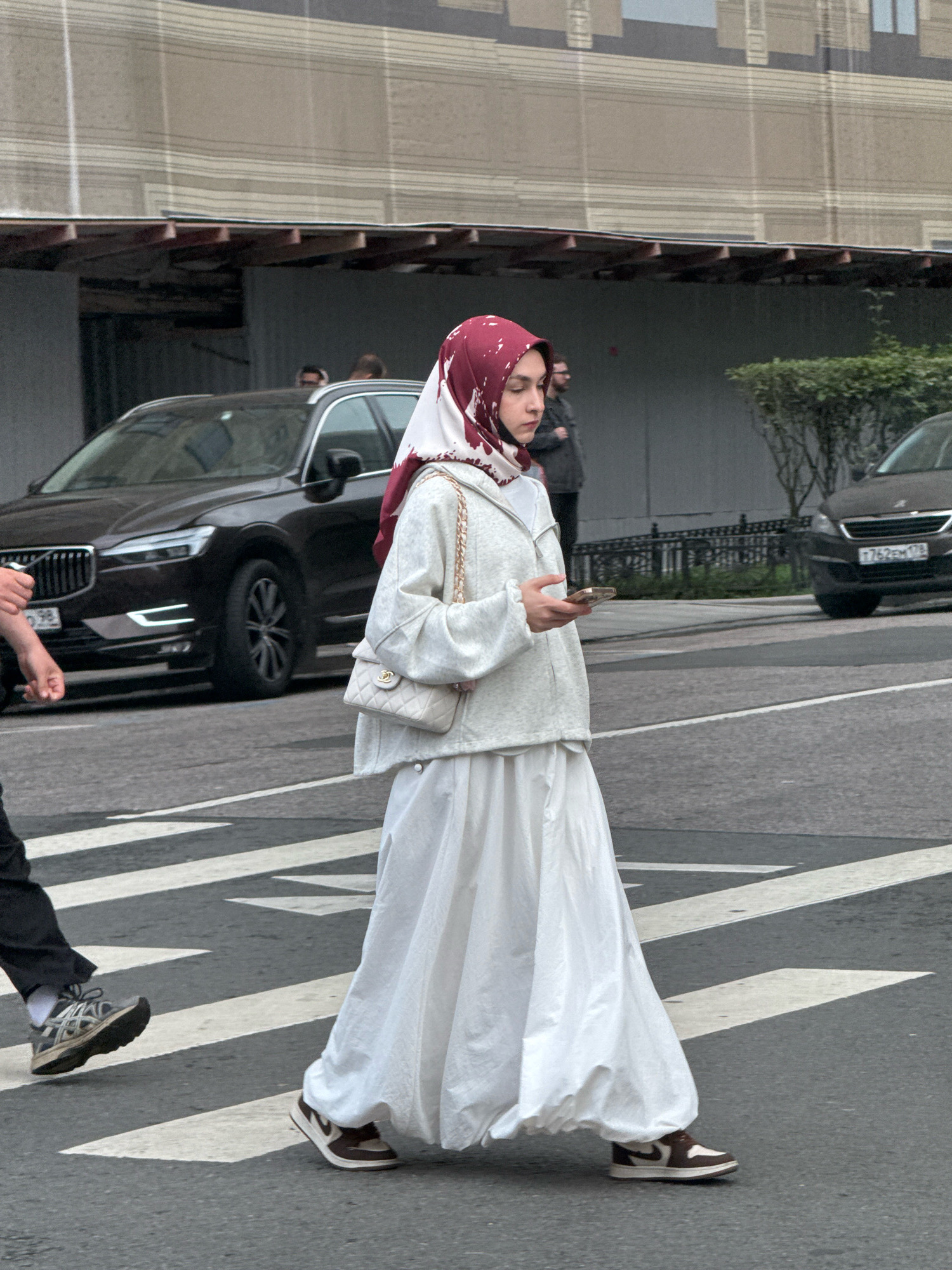 A young woman wearing a maroon and white hijab, a white oversized hoodie, and a long white skirt, walks across a pedestrian crossing. She is holding a mobile phone in her right hand. She is wearing sneakers. In the background, a dark-colored Volvo SUV is parked on the street, and a light-colored sedan is driving away. The woman is in Saint Petersburg, Russia, based on the license plates of the cars. The environment appears to be an urban street with a building facade in the background.