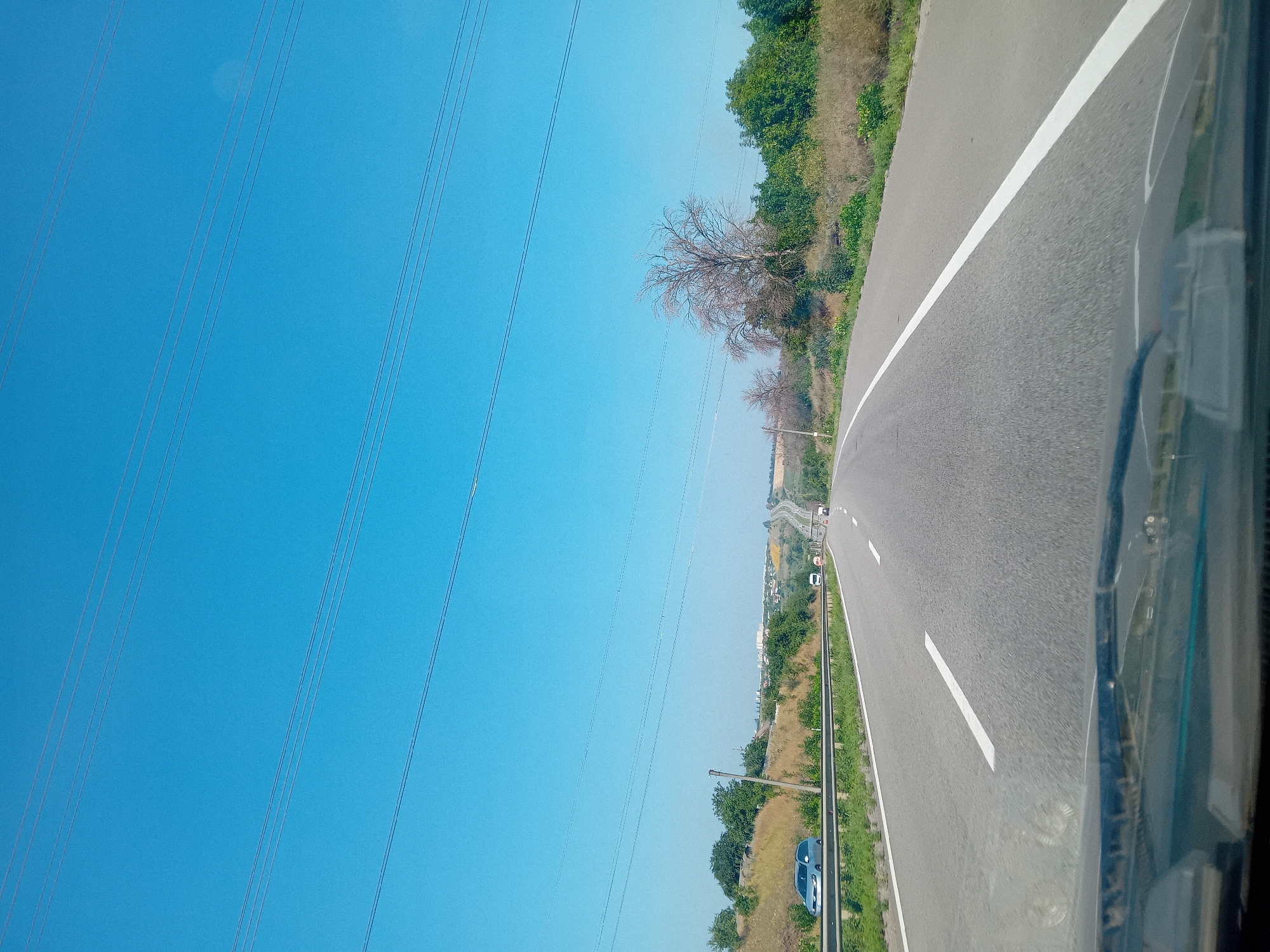 The image shows a view from inside a vehicle looking out at a highway in the city of Rohan, Ukraine. The sky is a clear blue with several power lines stretched across it. The highway is paved and has white lane markings. On the left side of the road, there is a metal guardrail and a grassy embankment with some trees and bushes. A silver car is driving on the highway. In the distance, there are more cars and some buildings. The overall scene depicts a typical highway landscape on a sunny day.