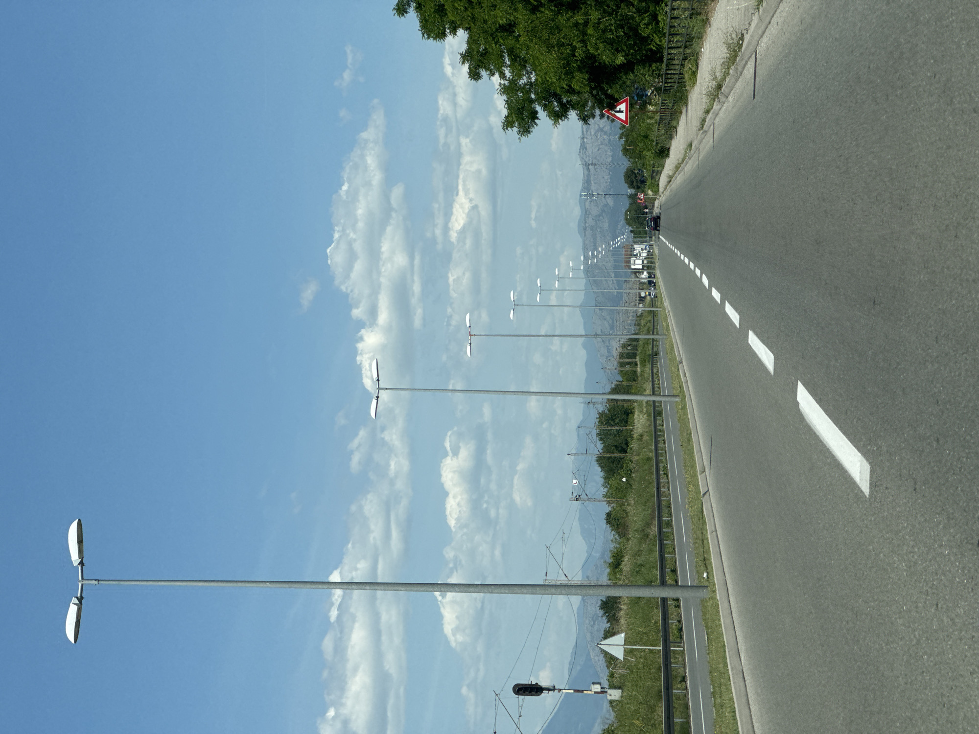 The image shows a straight, paved road in Mahala, Montenegro. The road has white dashed lines in the center and white solid lines on the sides. Several tall, silver light poles line the left side of the road. In the distance, vehicles can be seen on the road, along with power lines, mountains, and foliage. The sky is blue with scattered clouds. A "T" intersection sign is visible on the right.