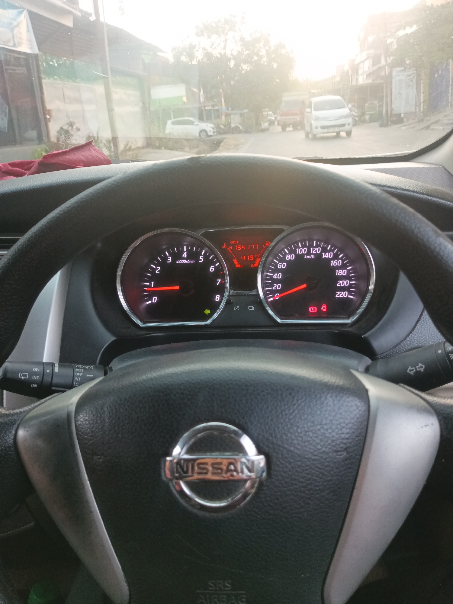 The image shows the interior of a Nissan car, focusing on the dashboard and steering wheel. The dashboard displays the tachometer and speedometer, with the engine RPM around 1000 and the car traveling at approximately 20 km/h. The odometer shows a reading of 154,177. Several warning lights are illuminated on the dashboard, including a brake warning light, an oil pressure warning light, and a battery warning light.
The steering wheel is black leather with a silver Nissan logo in the center. The turn signal and windshield wiper stalks are visible. Outside the car, a street scene is visible through the windshield. A white car is parked on the side of the road, and other vehicles are on the street. The lighting suggests it is daytime, possibly late afternoon or early morning due to the bright, hazy sunlight.
The image is taken from the driver's perspective, looking forward. The overall scene suggests the car is either stopped or moving slowly in a city or town environment. Based on visual cues and context, the location can be identified as Pangkajene, Indonesia.