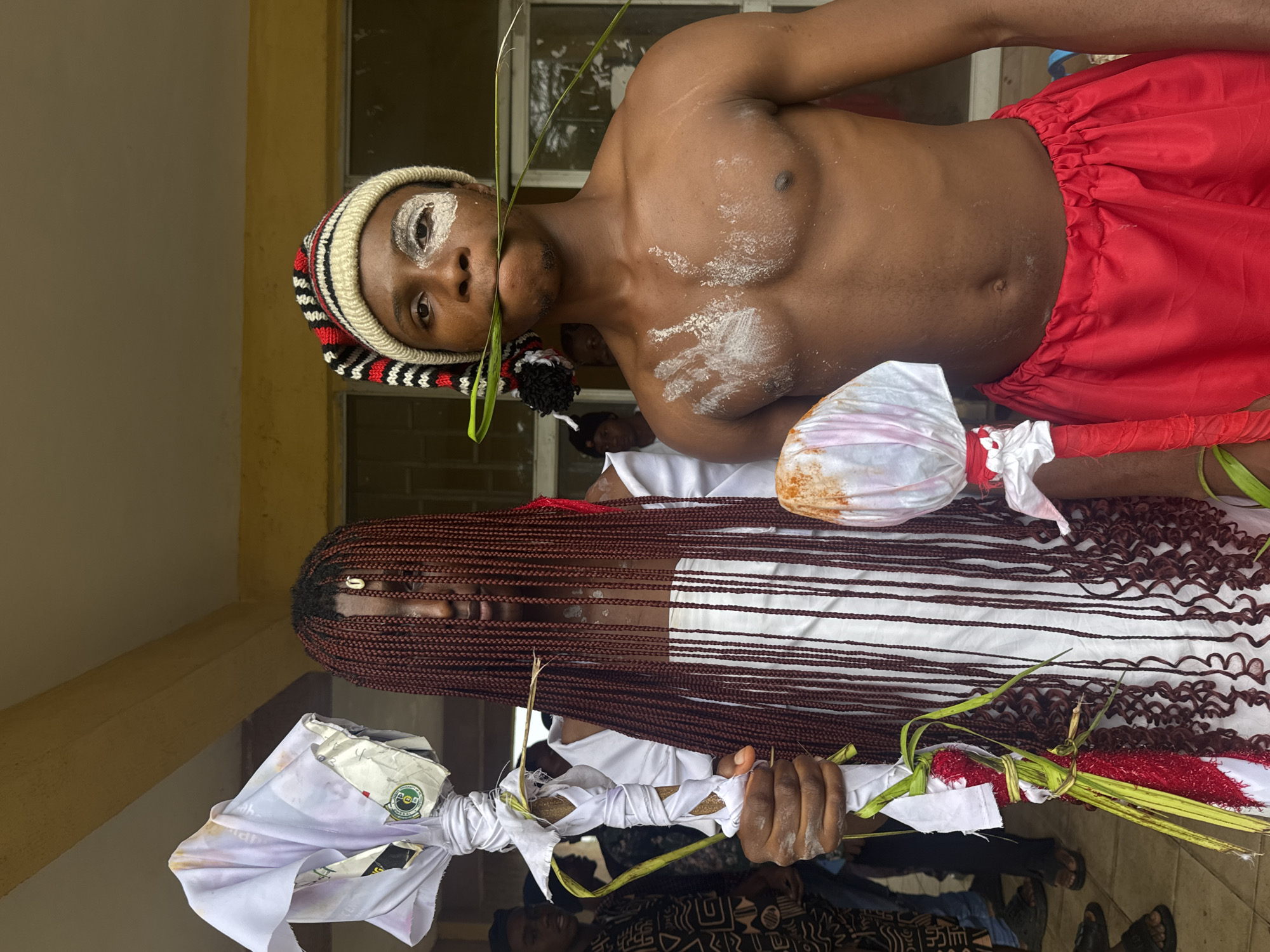 In Ihiagwa, Nigeria, two individuals stand prominently in the foreground. The person on the left, adorned with numerous dark, braided extensions, wears a white garment and clutches a staff wrapped in white fabric and adorned with green foliage. To the right, a bare-chested individual sports a colorful striped beanie, red shorts, and white markings on their face and chest, holding a green leaf in their mouth. Behind them, blurred figures and the exterior of a building with yellow walls and a beige ceiling are visible.