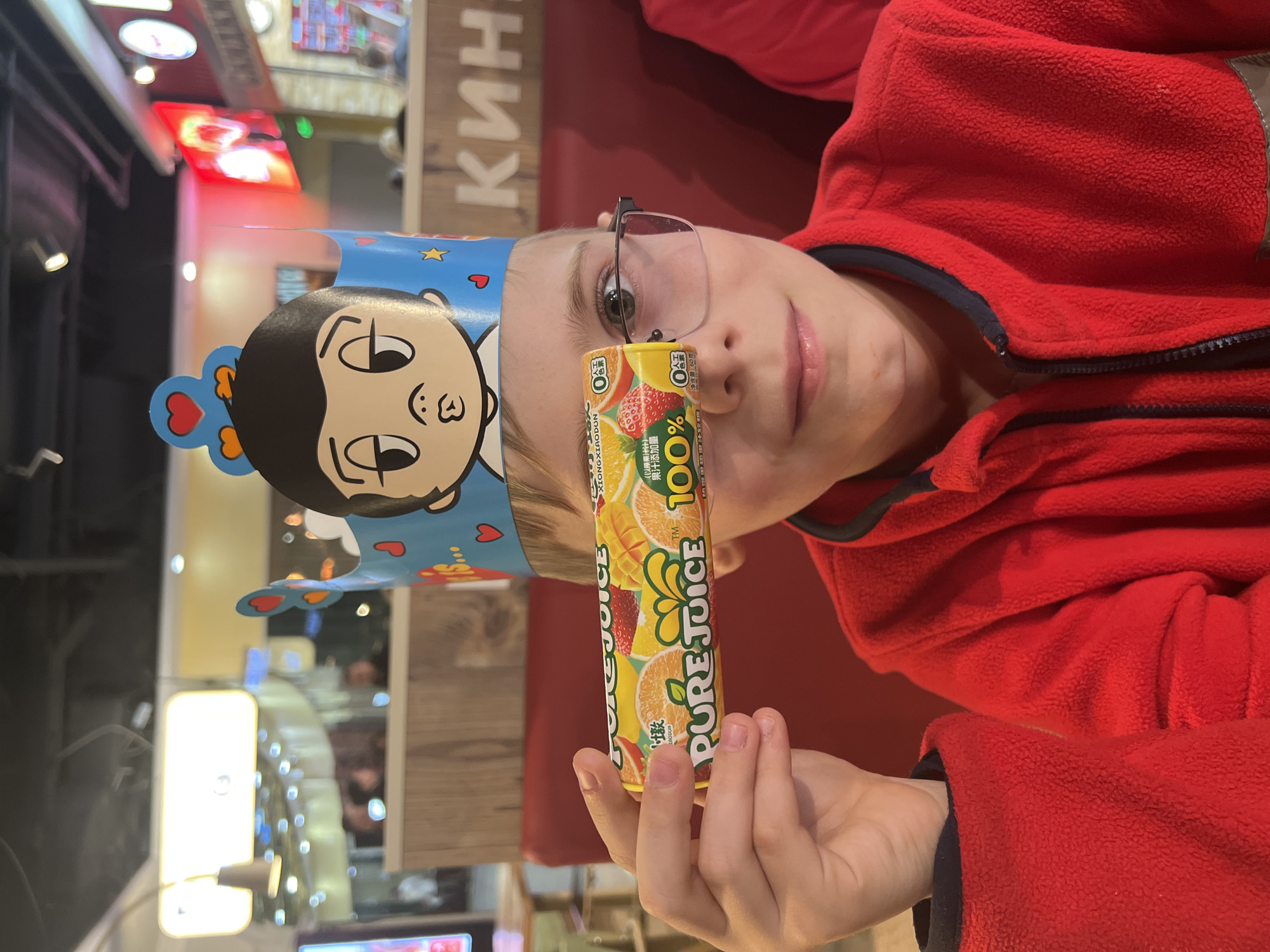 A young boy is depicted in an indoor commercial setting, identified as being in Yekaterinburg, Russia, likely a fast-food restaurant or food court. He is seated in a red upholstered booth, wearing a red fleece jacket with a black collar and zipper. On his head, he wears a blue paper crown adorned with hearts, stars, and a cartoon character featuring a prominent puckered mouth. The boy wears dark-framed glasses and holds a yellow rectangular candy bar labeled "PURE JUICE" horizontally over his left eye, partially obscuring it. The candy bar packaging displays images of oranges, strawberries, and mangoes, along with the text "100%" and various Chinese characters. He looks directly at the viewer with a slight smile.
The background is softly blurred but reveals elements of the environment. Behind the boy, light-colored wooden paneling is visible. To the viewer's left, a large, bright, illuminated sign or menu board is present. To the viewer's right, a prominent, illuminated sign displays the partial Cyrillic text "КИН", indicating a Russian location, possibly part of a larger sign such as "Burger King". Overhead, various light fixtures are visible, some casting bokeh effects. Other blurred figures of individuals and additional commercial signage are discernible further in the background, suggesting a busy public space.