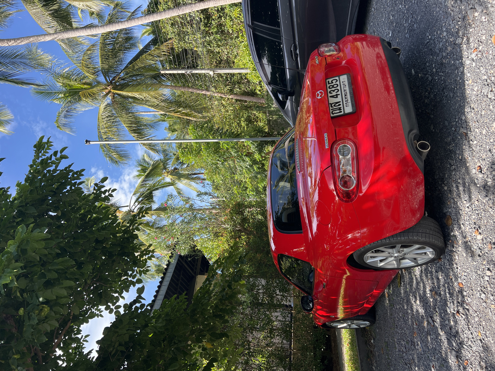 A red Mazda MX-5 is parked on a gravel lot in front of a building with a dark-tiled roof, surrounded by lush green foliage and tall palm trees under a clear blue sky with scattered white clouds. The car's rear is visible, featuring a license plate with Thai characters and the number "4385". Next to the red car, a dark-colored SUV is partially visible. Sunlight casts shadows from the trees onto the ground and the vehicles. The overall setting suggests a tropical or subtropical environment.