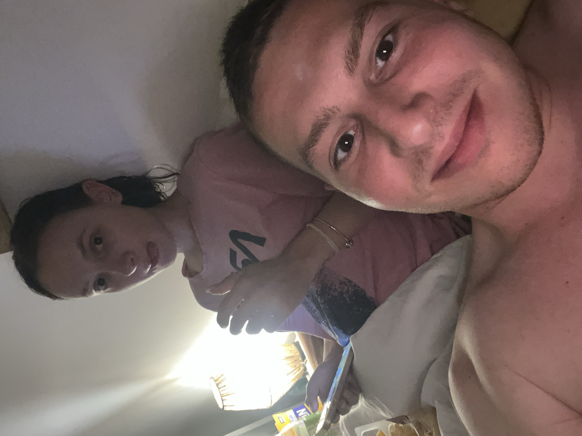 A selfie photograph shows two individuals indoors. A light-skinned male with short dark hair fills the right side of the frame, appearing closer to the camera. He has visible stubble and brown eyes, and appears to be shirtless. To the left and slightly behind him is a light-skinned female with long dark hair. She wears a pink t-shirt with a black graphic. She is holding a mobile phone. A bedside lamp illuminates the scene. In front of them, at the bottom of the frame, is food packaging. The location is unknown.