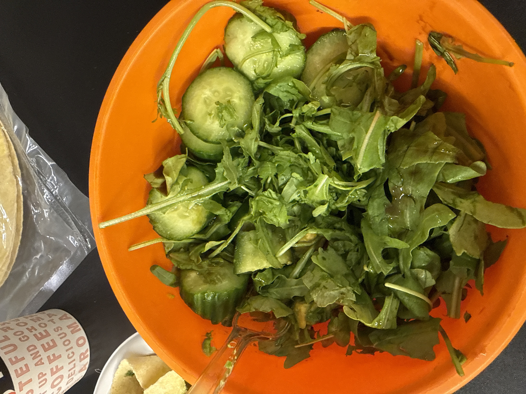 The image displays an overhead view of a fresh green salad served in a vibrant orange bowl, set against a dark, possibly black, surface. The salad is composed mainly of arugula leaves and several round slices of cucumber, appearing moist, possibly from a dressing. A clear plastic serving utensil, resembling a pair of tongs, is partially submerged in the salad.
To the left of the orange bowl, a stack of light-colored, round flatbreads or tortillas is visible, sealed within a transparent plastic bag. Below the tortillas, a white paper cup with brown text is partially in view. Recognizable words on the cup include "FUL," "COFFEE," "DELICIOUS," "UP AND GO," and "ROMA," indicating it is likely a branded coffee cup. A small, white rectangular dish containing several small, cubed, light-colored food items is partially visible at the bottom right corner of the frame.
The scene suggests an indoor setting, perhaps a kitchen counter, dining table, or office break area, where food is being prepared or served. There are no people present, and no specific activity beyond the presentation of food is clearly taking place. The time of day and weather are not discernible. No specific visual cues in the image indicate the provided location context of Tel-Aviv, Israel.