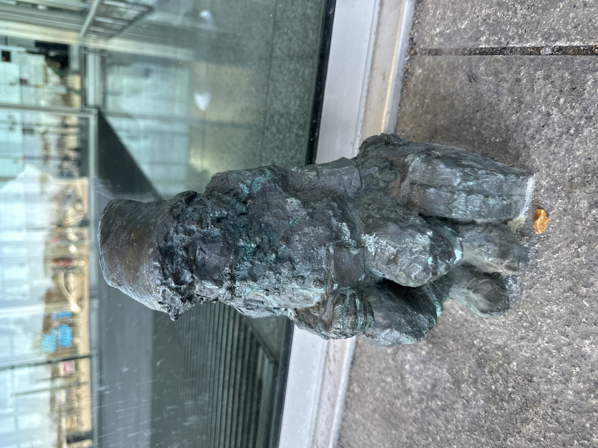 The image shows a bronze sculpture of a dwarf, one of the numerous "Krasnale" (dwarfs) found in Wrocław, Poland. The sculpture is located on a paved surface, likely a sidewalk, adjacent to a modern building with glass windows, which reflect the surrounding urban landscape. The dwarf sculpture is patinated with green and dark gray tones. A small, yellow object is visible near its base on the pavement.
