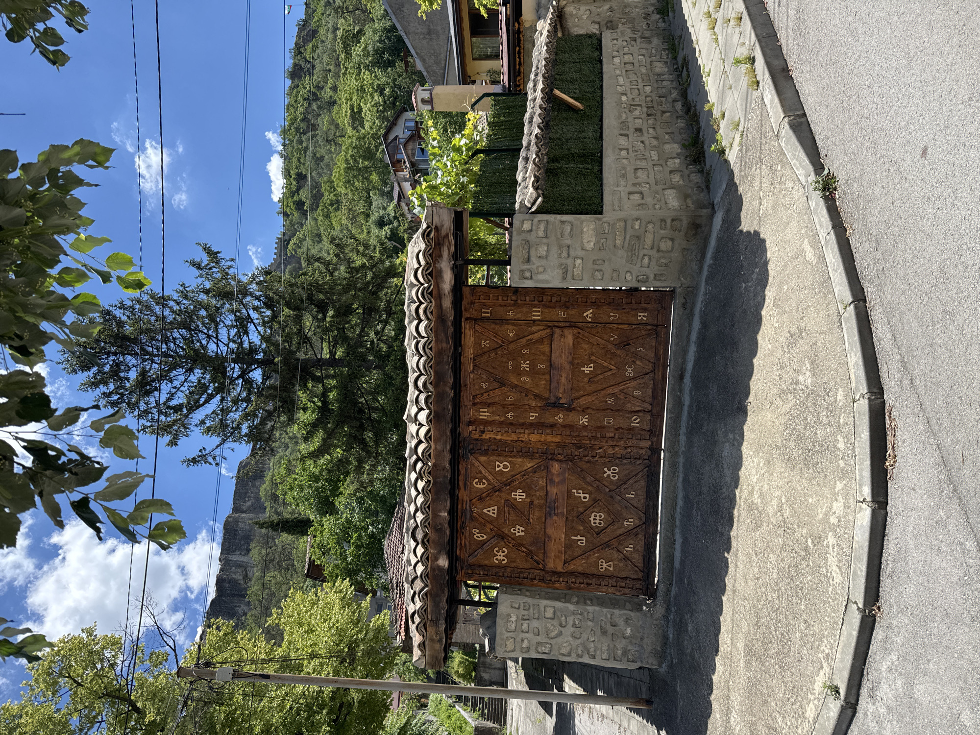 The image shows a large, ornate wooden gate in the city of Provadia, Bulgaria. The gate is decorated with intricate carvings that appear to be of Cyrillic letters or symbols. The gate is set within a stone wall and is topped with a tiled roof. Behind the gate, there is a lush green landscape with trees and houses on a hillside. The sky is blue with scattered clouds. The foreground shows a paved area with a curb and a street. The overall scene depicts a traditional architectural style in a mountainous or hilly urban environment.