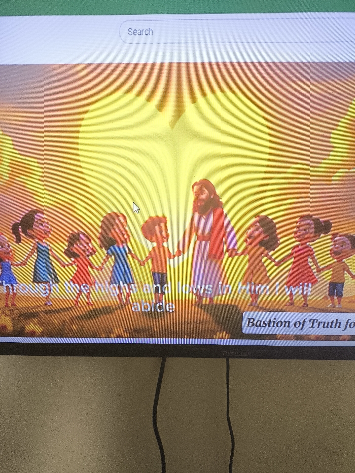 The photograph was captured in Nicosia, Northern Cyprus / Cyprus. It depicts a digital screen, likely a television or monitor, displaying an animated scene. The screen shows a central bearded figure, resembling Jesus Christ, wearing a striped robe, holding hands with approximately eight diverse cartoon-style children arranged in a semi-circle. They are positioned on a light brown surface against a radiant yellow and orange background with emanating light rays. Visible text overlays include "through the highs and lows in Him will abide" at the bottom center, and "Bastion of Truth for" within a rectangular box at the bottom right. A white "Search" bar is present at the top of the screen, and a white arrow mouse cursor points towards the center-left. Moiré patterns are visible across the screen surface. Below the screen, a textured, light beige wall is visible, with two black cables hanging vertically downwards.
