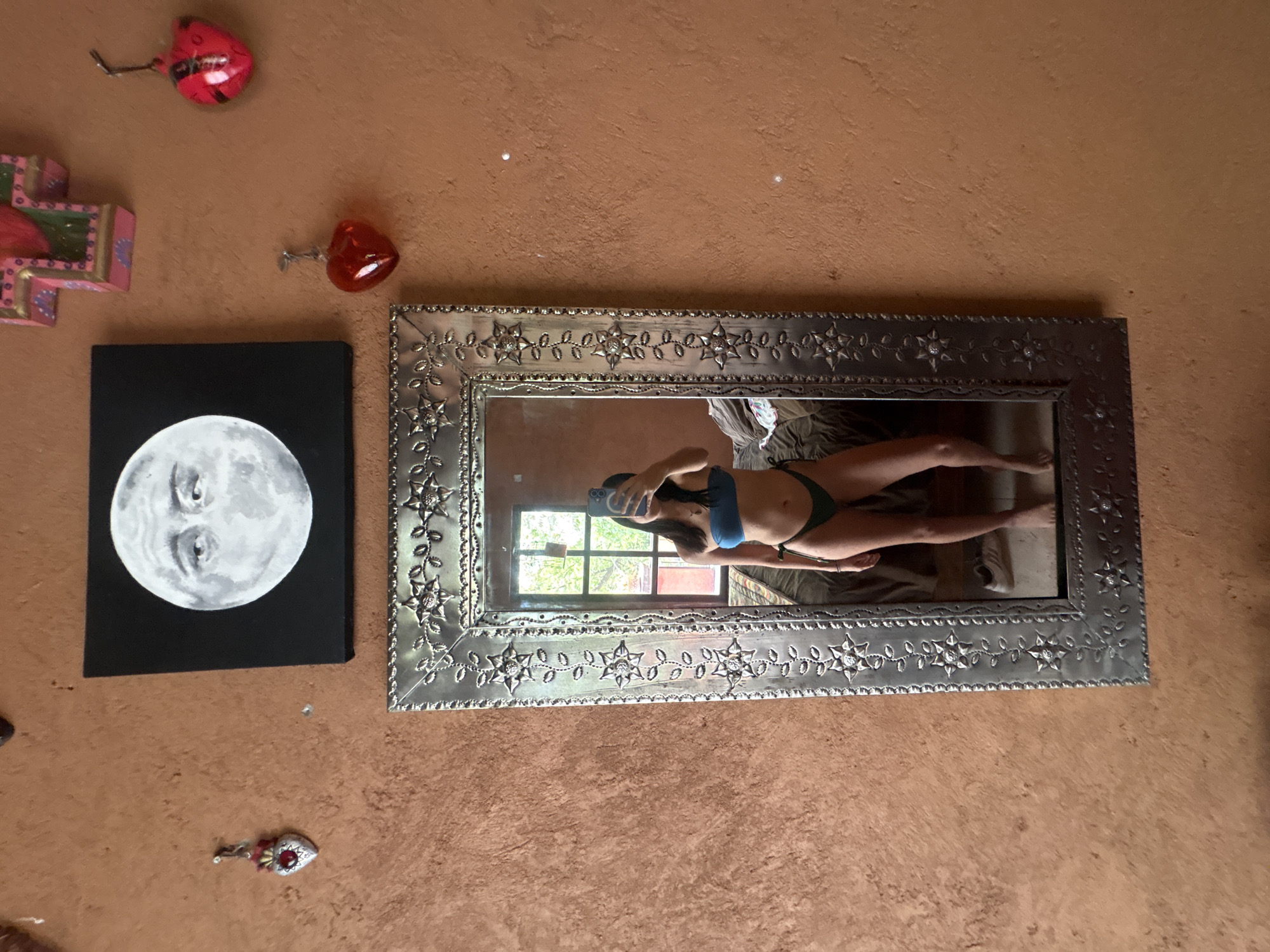 The scene is an interior wall of a building in Cañada de Madero, Mexico. The wall, finished in a rough orange stucco texture, features a rectangular metal-framed mirror reflecting a person wearing a blue top and green bottoms. The person is holding a smartphone, presumably taking the picture. The reflection also shows a window, greenery outside, and part of a bed or seating area. To the left of the mirror is a black square canvas with a white painting of a moon-like sphere with eyes. Above the mirror and moon painting are decorative items: one is a red heart-shaped object, and another is a painted wooden cross. There's also a ladybug-shaped object attached to the wall above the cross. At the bottom of the wall, near the moon painting, there is a decorative ornament.
