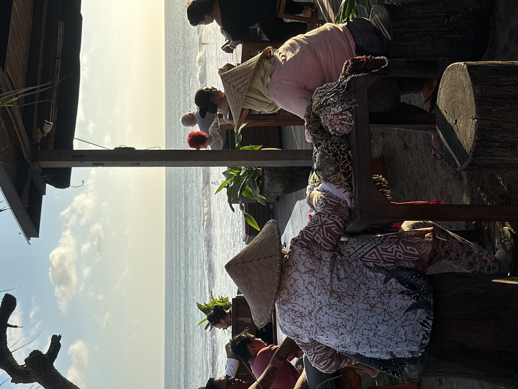 The image depicts a scene at a seaside establishment in North Kuta, Indonesia. Several people are present, seated at wooden tables and chairs crafted from tree trunks and branches. Some are wearing traditional conical hats. The sky is bright, with a horizon view of the ocean. The establishment appears to be partially covered by a wooden structure.