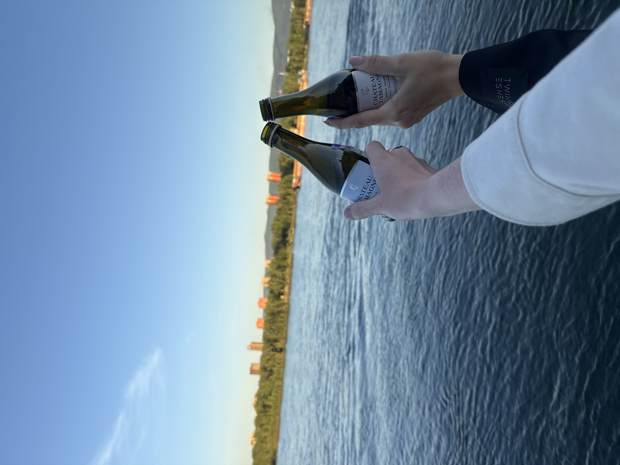 The image captures two people's hands holding small bottles of sparkling wine or champagne over a body of water, possibly a river or lake, under a clear blue sky. The photo is taken in a portrait orientation but is rotated sideways.
The subject consists of two hands, extending forward. The left hand, appearing more masculine, has a ring on its index finger and holds a bottle with a label visible as "CHATEAU TAMAGNE BRUT PURE". The right hand, appearing more feminine with light-colored fingernails, holds a similar bottle labeled "CHATEAU TAMAGNE BRUT ROSÉ". A black sleeve on the right arm features a tag with the word "TWINSENSE" printed vertically.
The setting is outdoors, overlooking a vast expanse of dark blue water with subtle ripples. In the distant background, a shoreline is visible, densely covered with green trees and several light-colored buildings scattered among them. The sky is bright and clear, displaying a gradient from a darker blue overhead to a lighter, almost white-blue, near the horizon.
The activity depicted appears to be a toast or a celebratory moment, with the bottles held out. The clear sky and bright lighting suggest it is daytime, possibly late afternoon or early evening. The overall mood is serene and celebratory. Given the context, this scene is likely taking place in Krasnoyarsk, Russia.