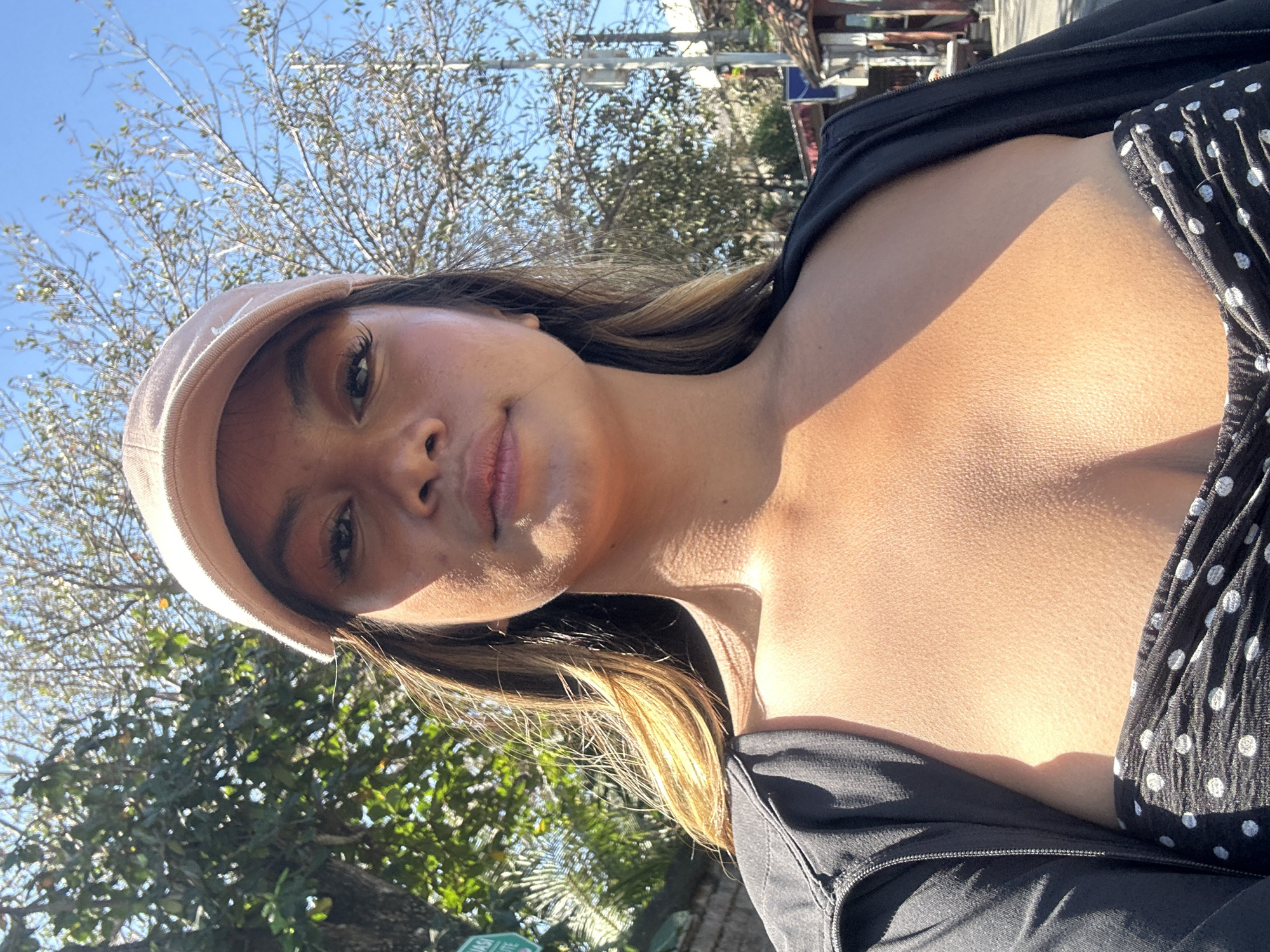 A woman wearing a beige baseball cap and a black zip-up jacket over a black and white polka dot top is photographed from the chest up, outdoors. Sunlight casts a shadow across her upper body. In the background, trees with sparse foliage and a bright blue sky are visible, suggesting a warm, sunny day. A street sign with green lettering can be seen on the left side of the image. The overall scene appears to be in a tropical or subtropical urban environment.