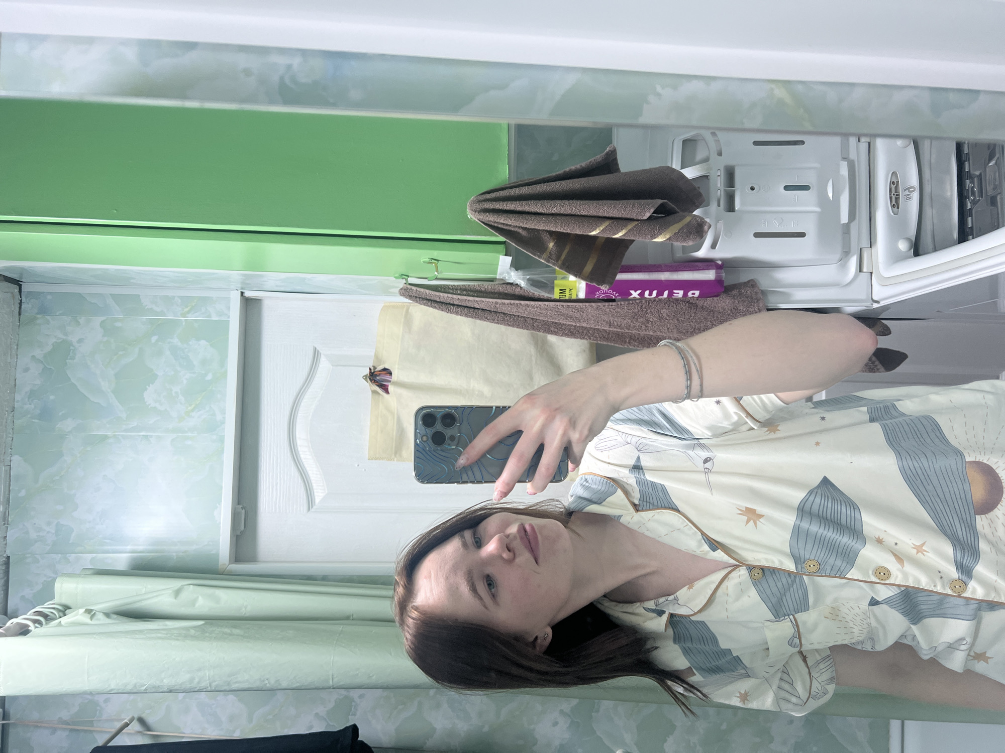 An indoor mirror selfie depicts a young woman with long, dark brown hair, wearing light-colored pajamas adorned with patterns of foliage, stars, and what appears to be a sun or moon. She holds a smartphone (an iPhone with multiple cameras) and looks directly into the camera with a neutral, slightly pleasant expression. The image is taken in what appears to be a bathroom or utility room, likely in a private residence in Chaikovsky, Russia.
The background, visible in the mirror's reflection, features a light green or mint-colored textured wall covering. Above the mirror, a bright green shelf or cabinet is reflected. To the right, a folded brown towel with gold stripes rests near a white plastic compartment, possibly a detergent dispenser from a washing machine or a similar appliance, which shows various indentations and symbols. A purple package, partially visible, has the word "DETOX" written on it (reversed due to the mirror reflection). Further back, a white door with decorative molding can be seen, along with a light brown paper bag or similar item. The overall lighting suggests an indoor setting, and no specific time of day or weather conditions are discernible from the image.
